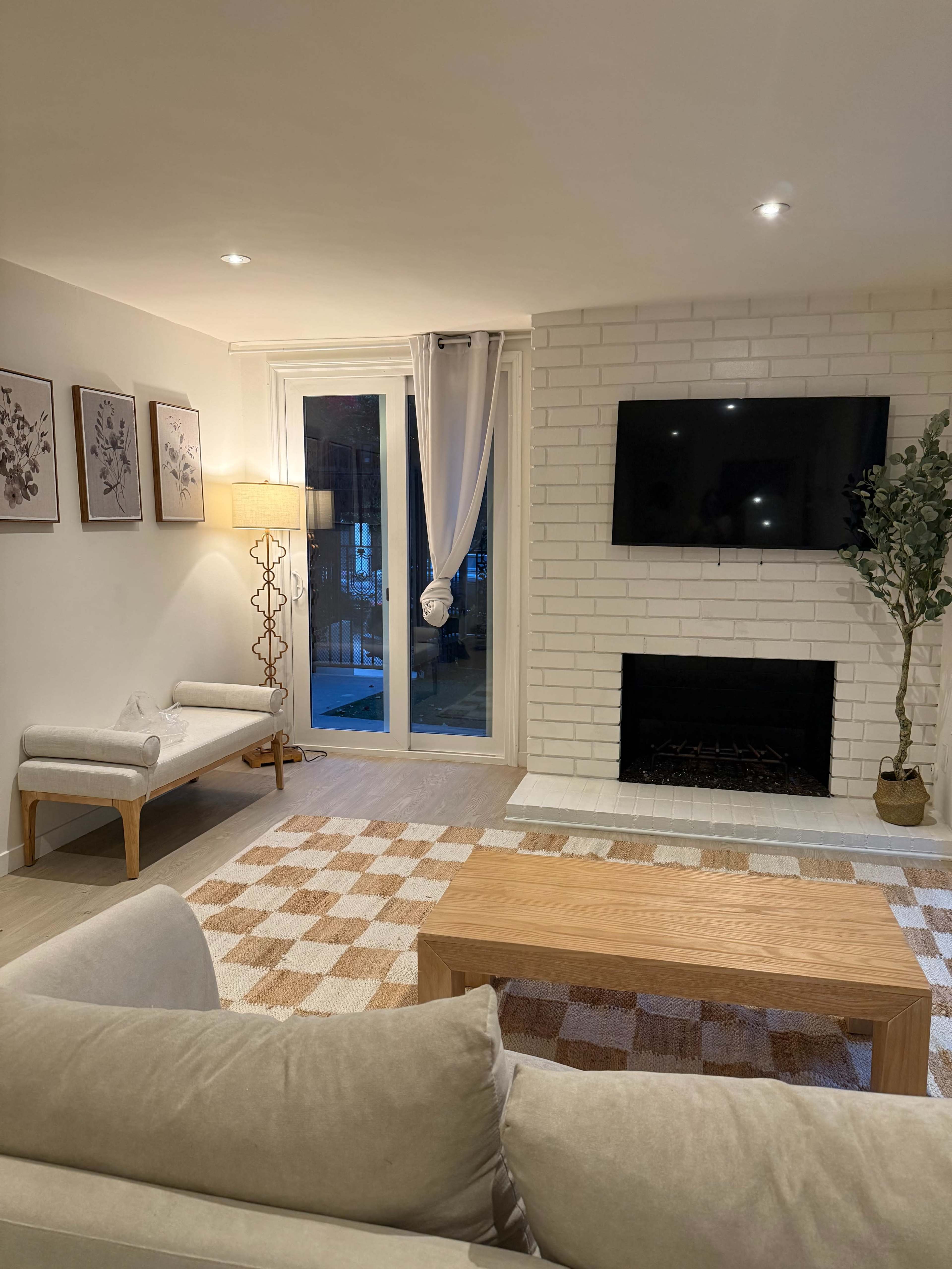 The image shows a modern living room with a white brick fireplace, a television mounted above it, a wooden coffee table, a light-colored sofa, and artwork on the walls.