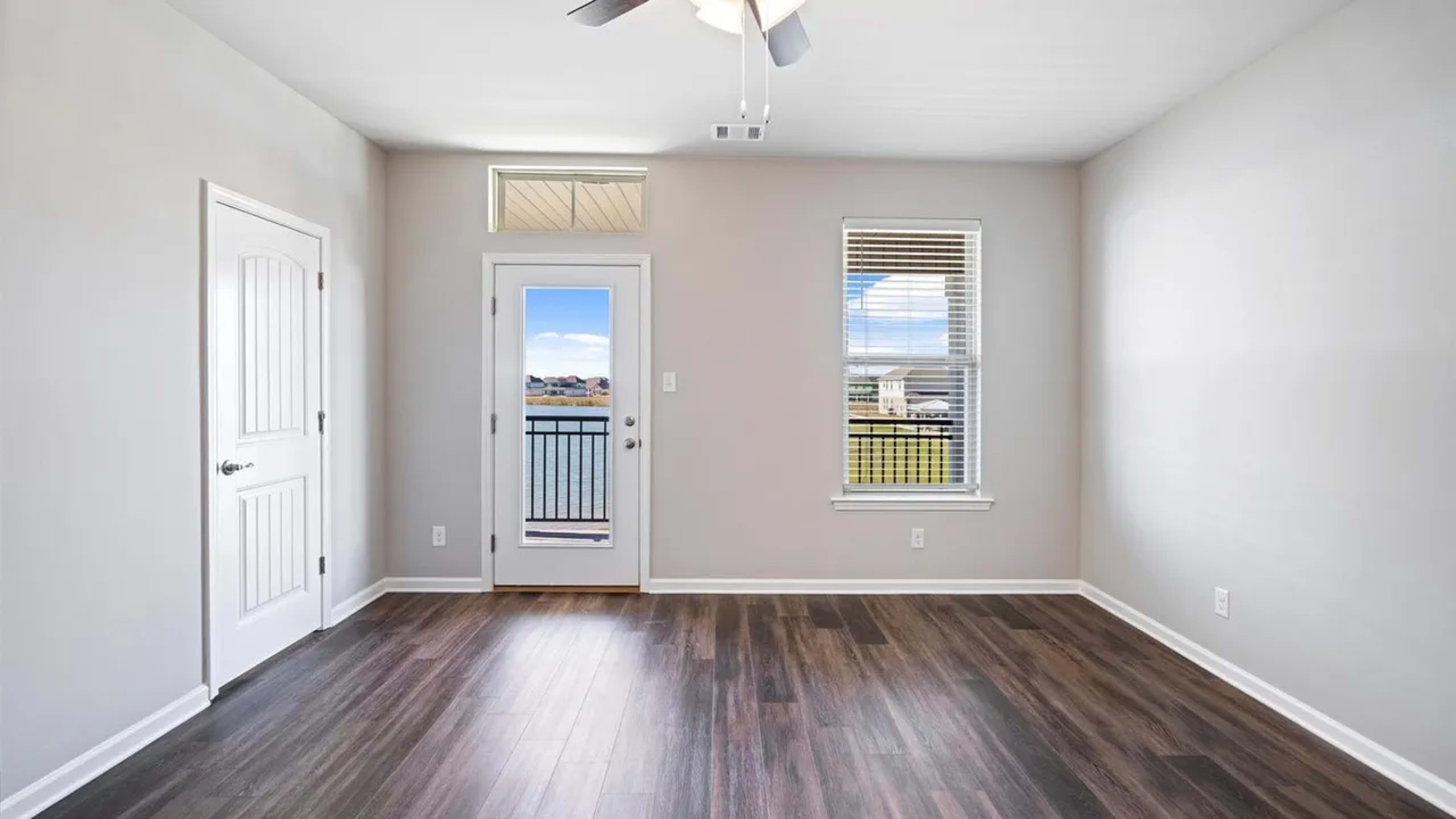 The image shows a minimalist room with hardwood floors, a ceiling fan, and a door that leads to a balcony with a view.