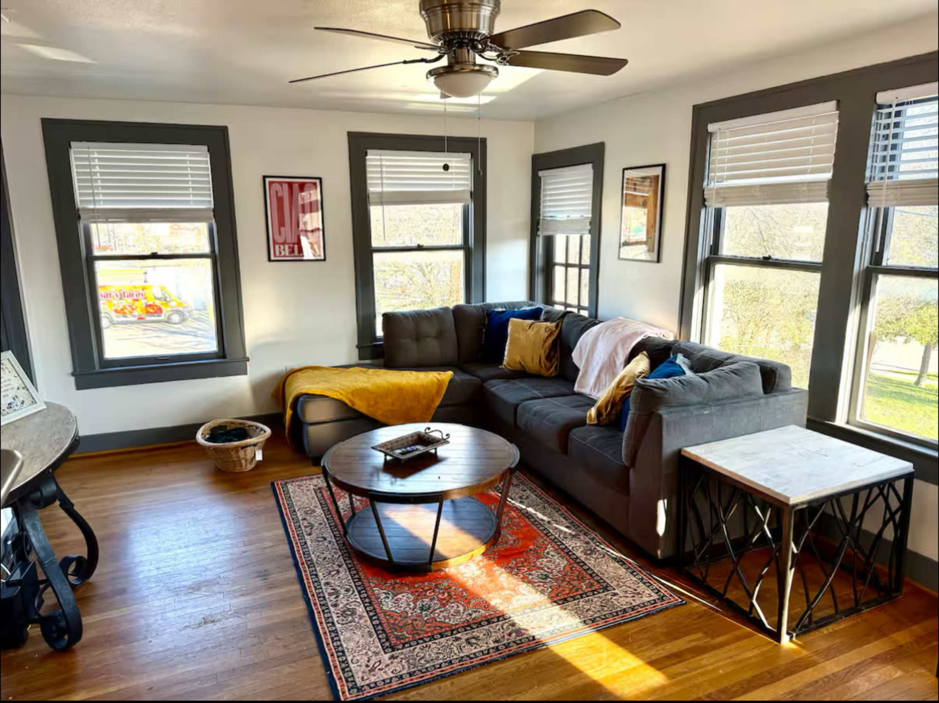 The image shows a cozy living room with a gray sectional sofa, a round coffee table, and large windows allowing natural light to fill the space.