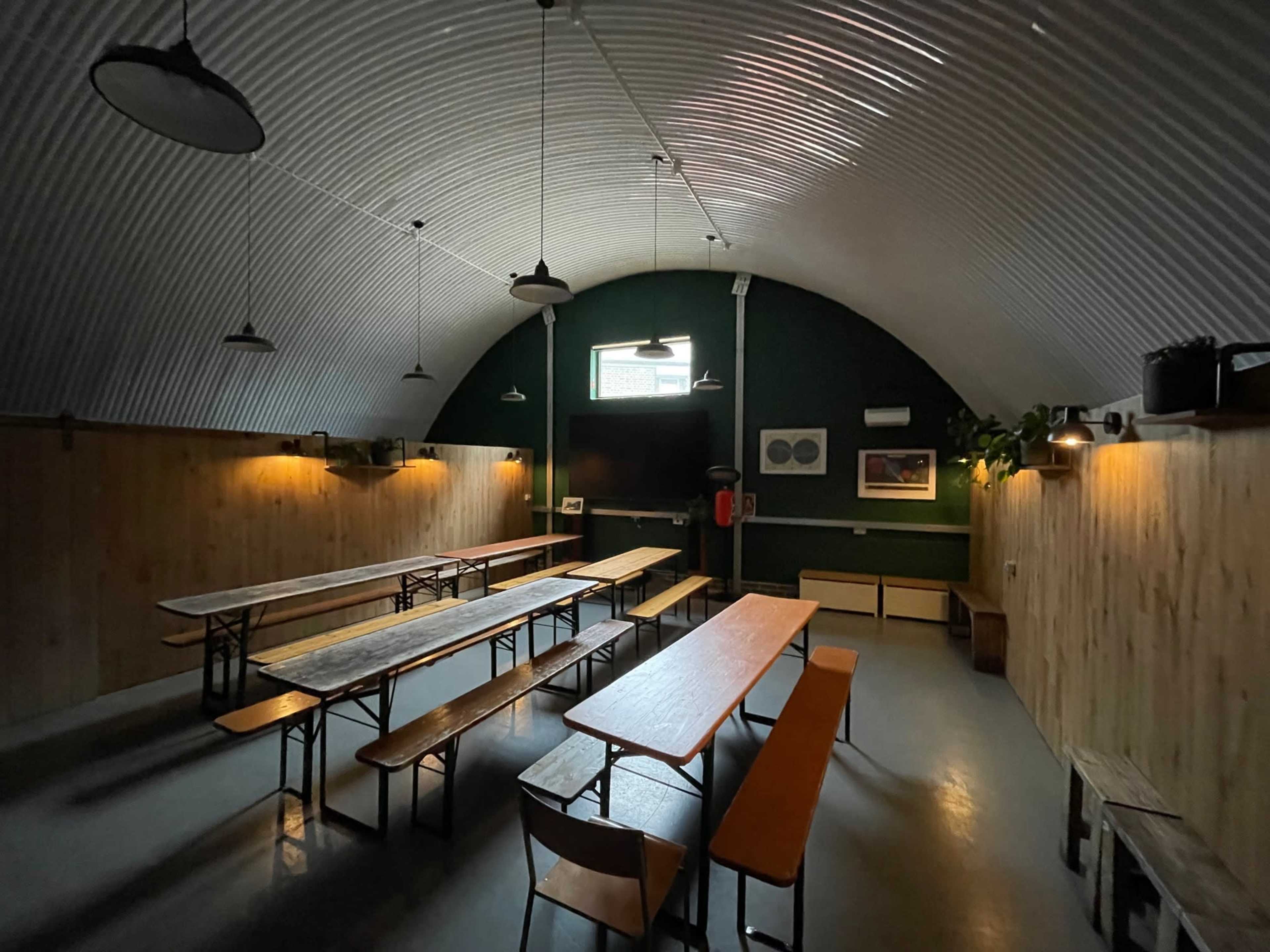 The image depicts a spacious, dimly lit room with wooden tables and benches arranged in rows, featuring a flat-screen TV at the front and plants on the walls.