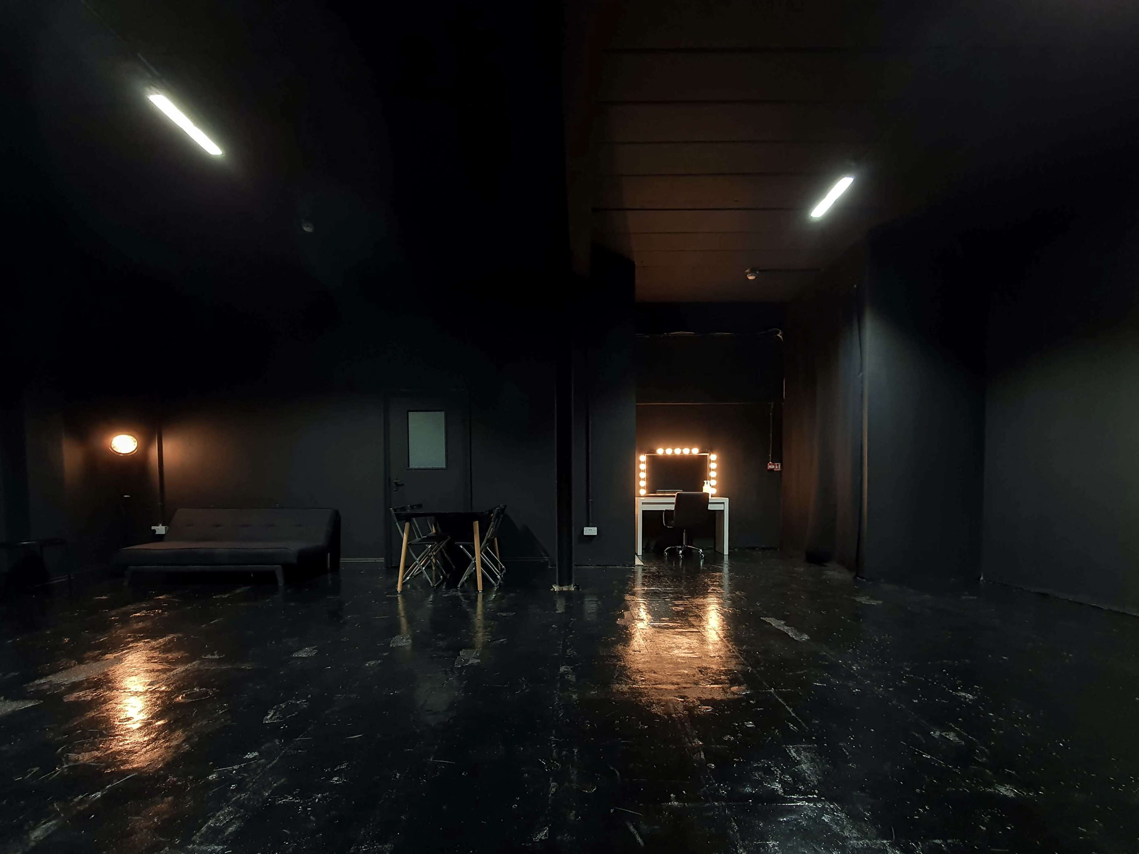 A dimly lit room with black walls and a shiny floor, featuring a sofa, a table, and a vanity with illuminated mirrors at the far end.