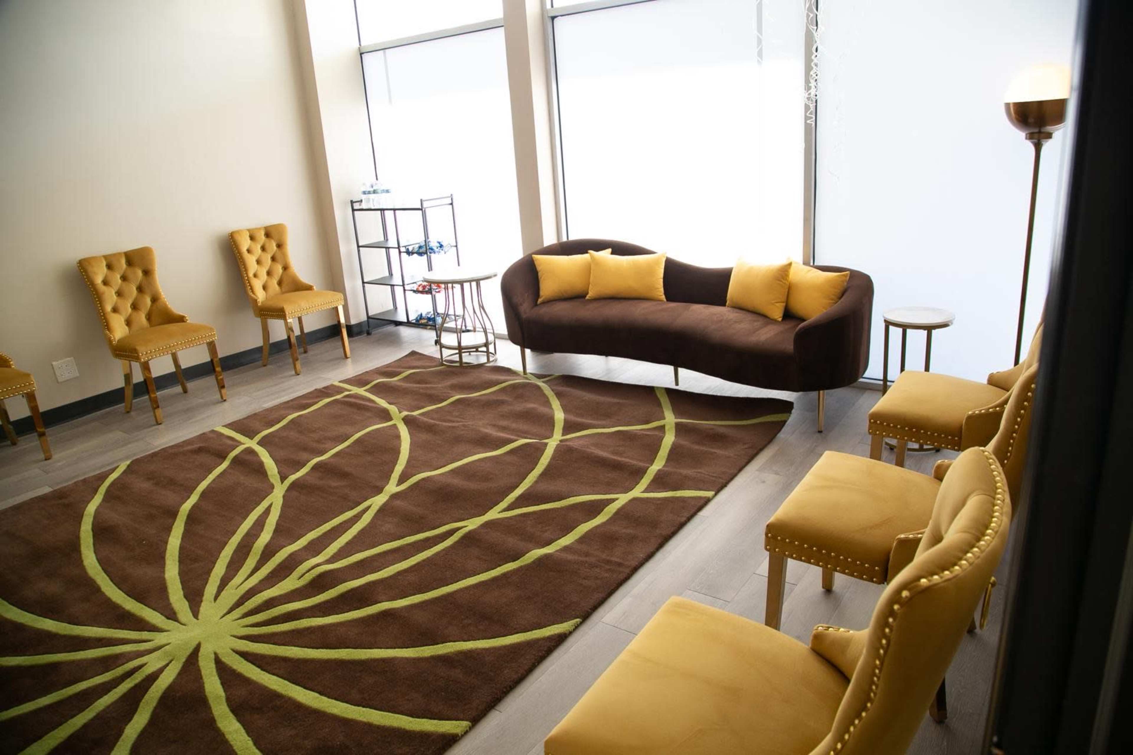 A modern waiting area features a brown curved sofa, yellow accent chairs, and a large rug with a floral design.