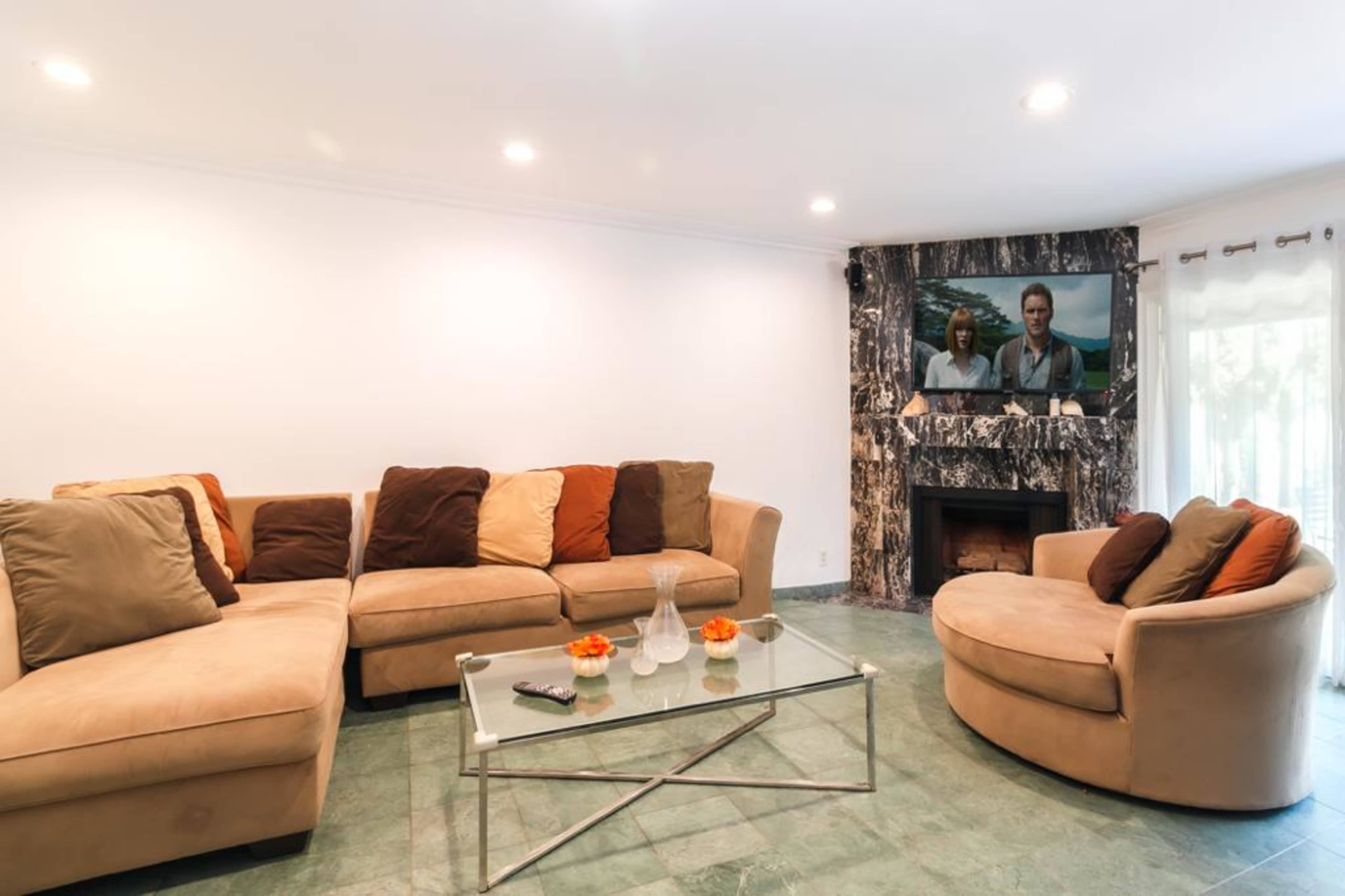 A spacious living room features a large sectional sofa, a round chair, a glass coffee table, and a TV mounted above a stone fireplace.