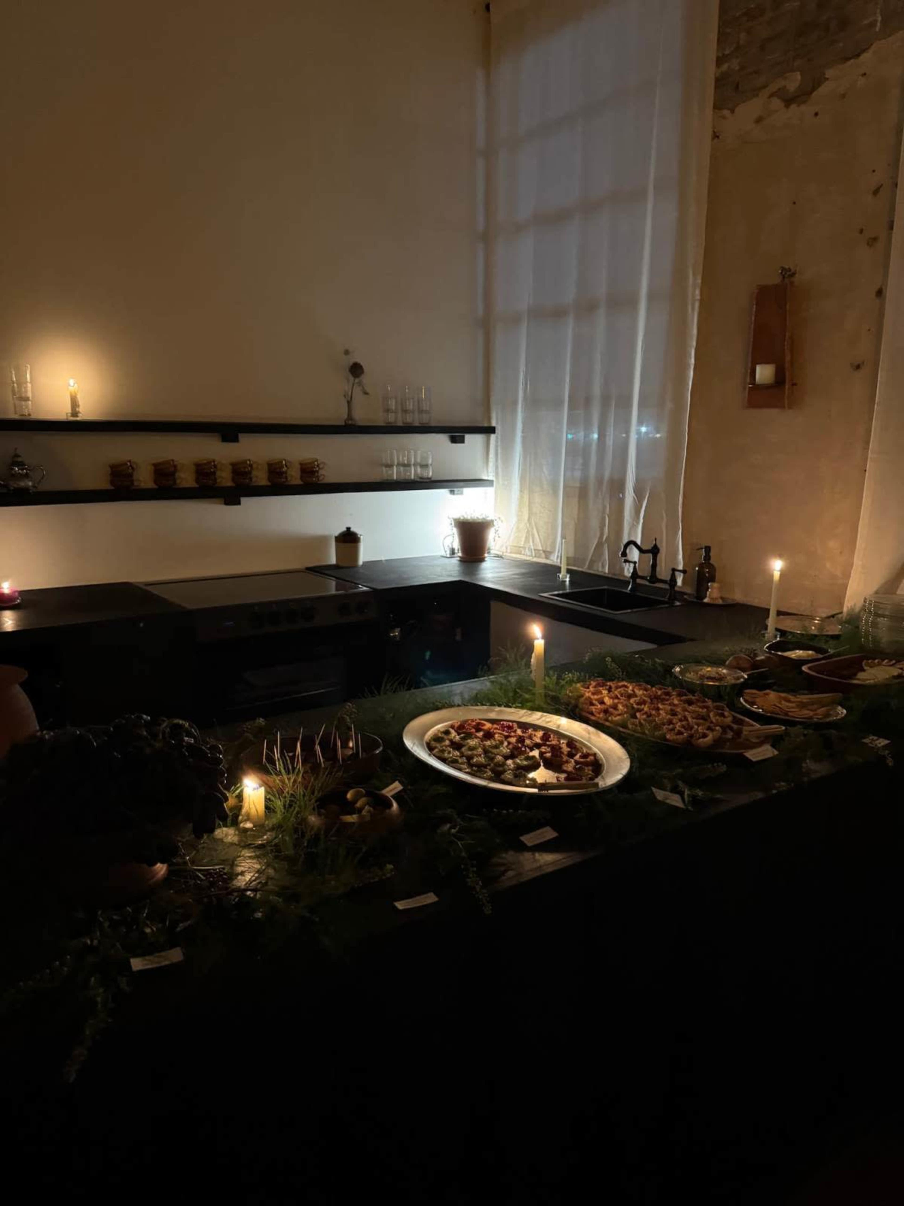 A dimly lit kitchen scene features a wooden table adorned with an array of elegantly arranged dishes and candles, surrounded by greenery and shelves holding jars.