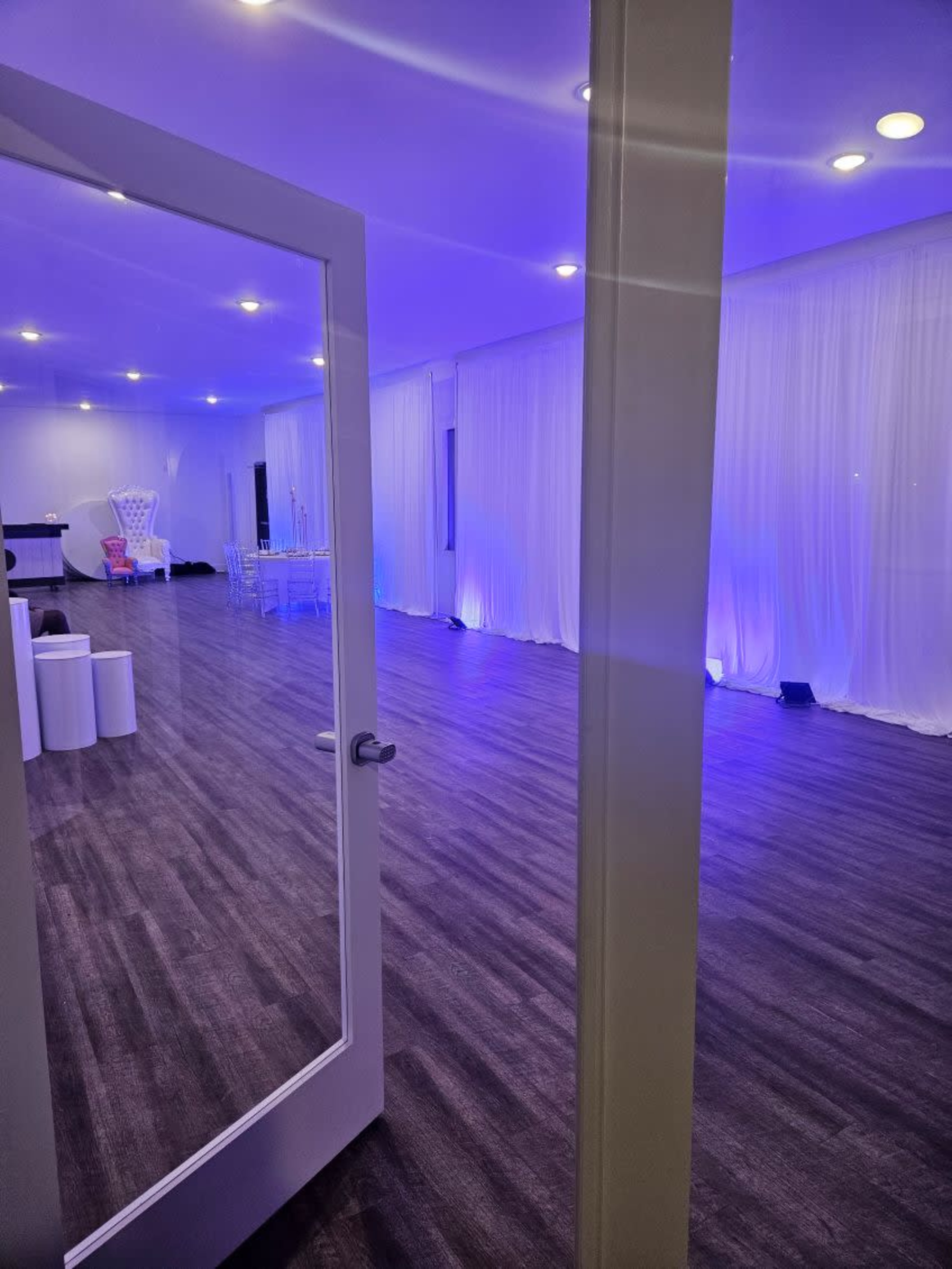 The image shows a spacious interior room with wooden flooring, white drapes, and blue lighting.