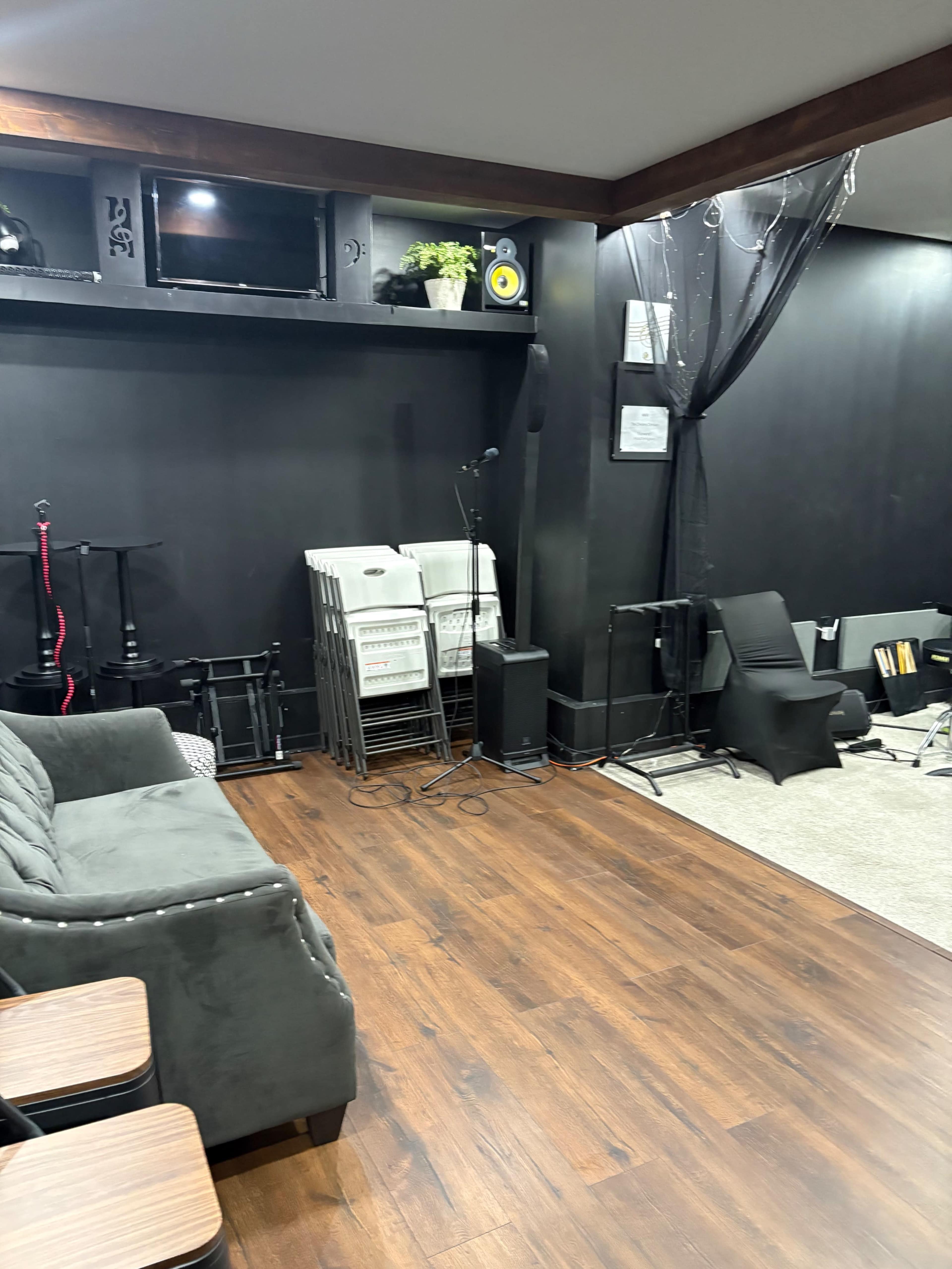 The image shows a dark-colored room with a gray sofa, a microphone stand, and various music equipment arranged along the walls.