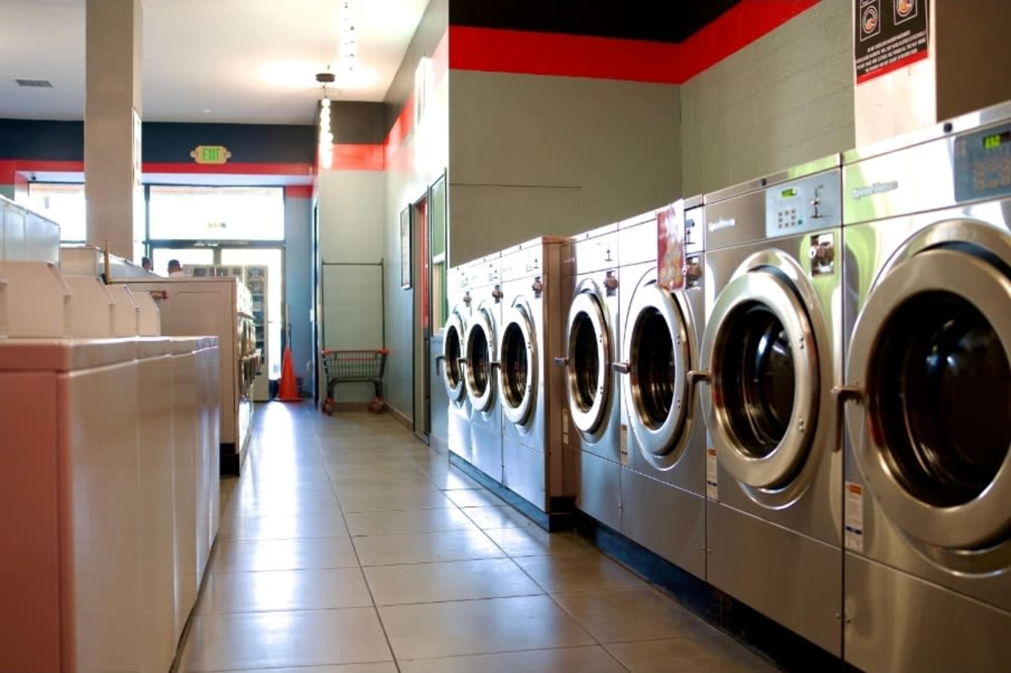 The image shows a row of front-loading washing machines in a laundromat, with a bright, clean interior and a shopping cart visible in the distance.