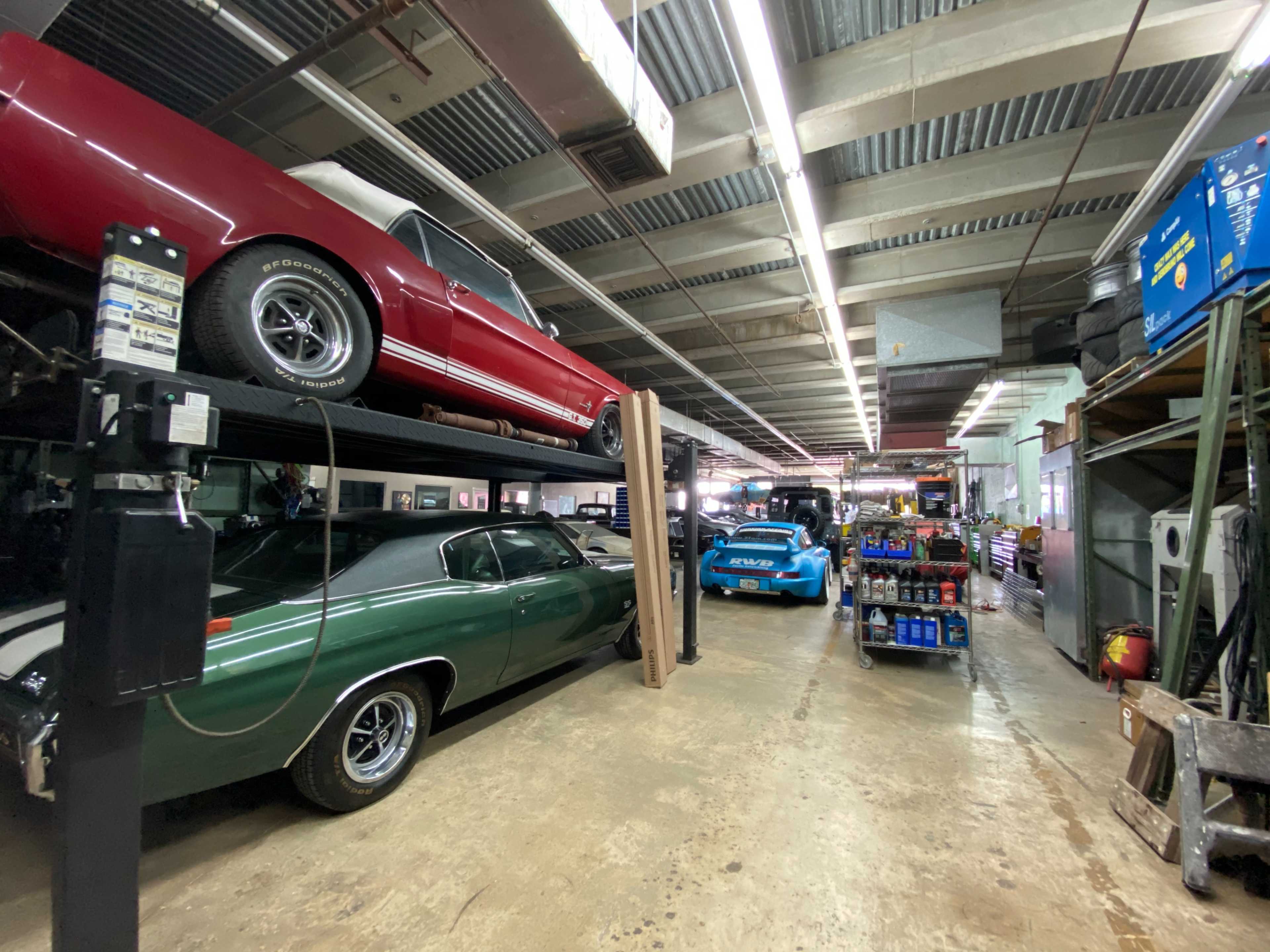 The image shows a car workshop with several vehicles, including a green car on the lower level and a red convertible on a lift, along with tools and equipment lined along the workspace.
