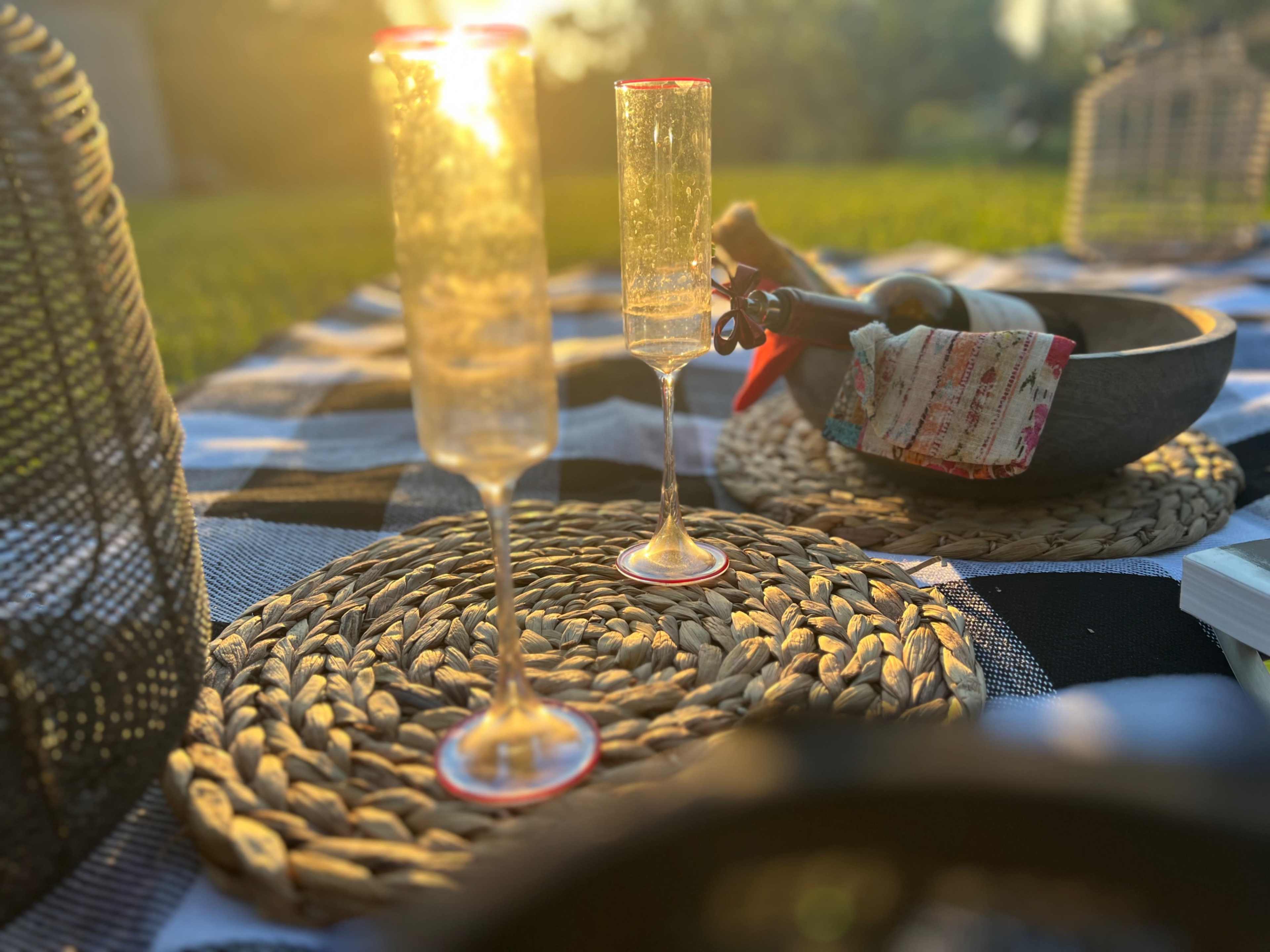 The image depicts two champagne flutes on woven placemats, with a sunlit picnic setting featuring a bowl and decorative items in the background.