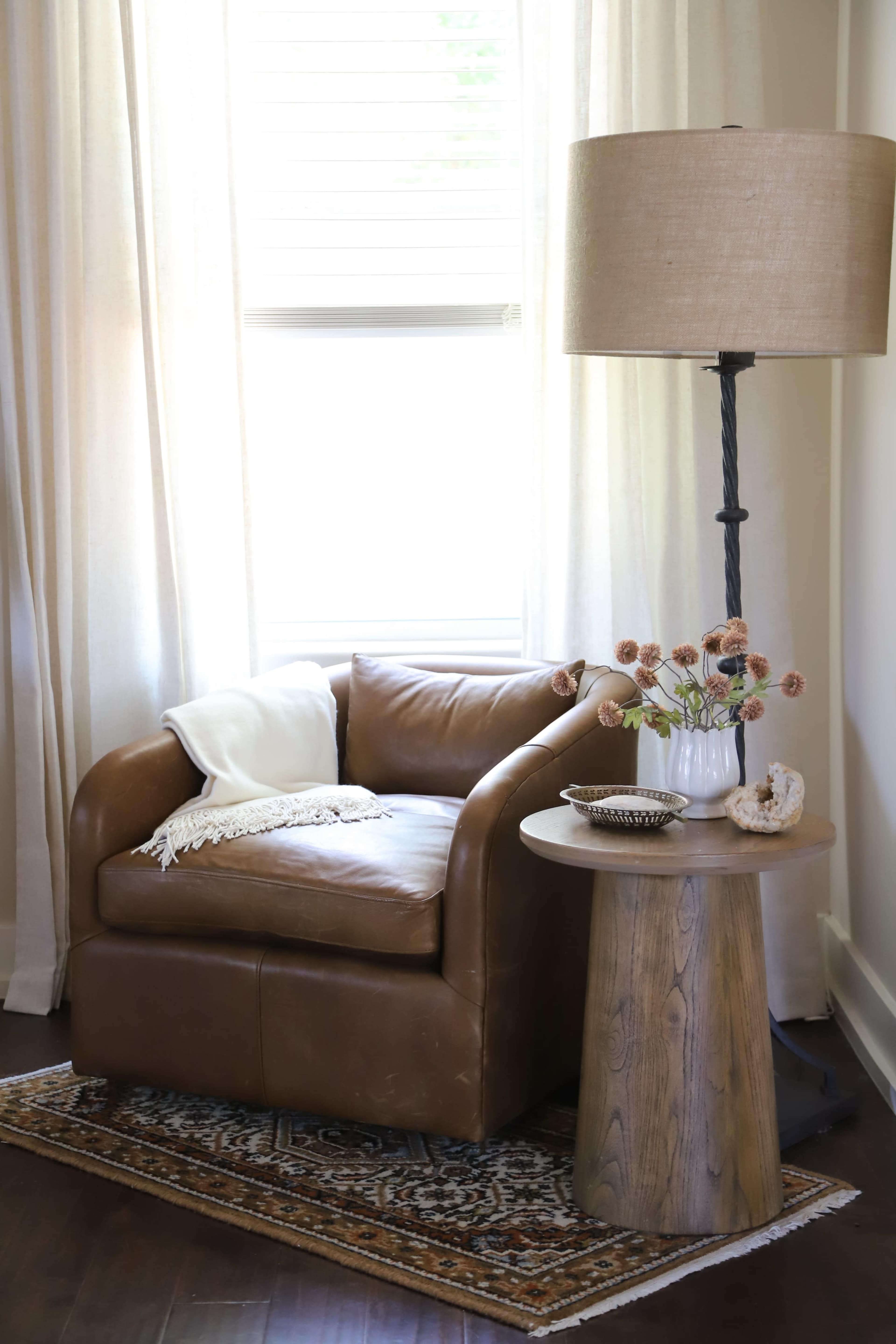 A brown leather armchair is positioned next to a round wooden side table with a lamp and a vase of dried flowers, located near a window with light curtains.