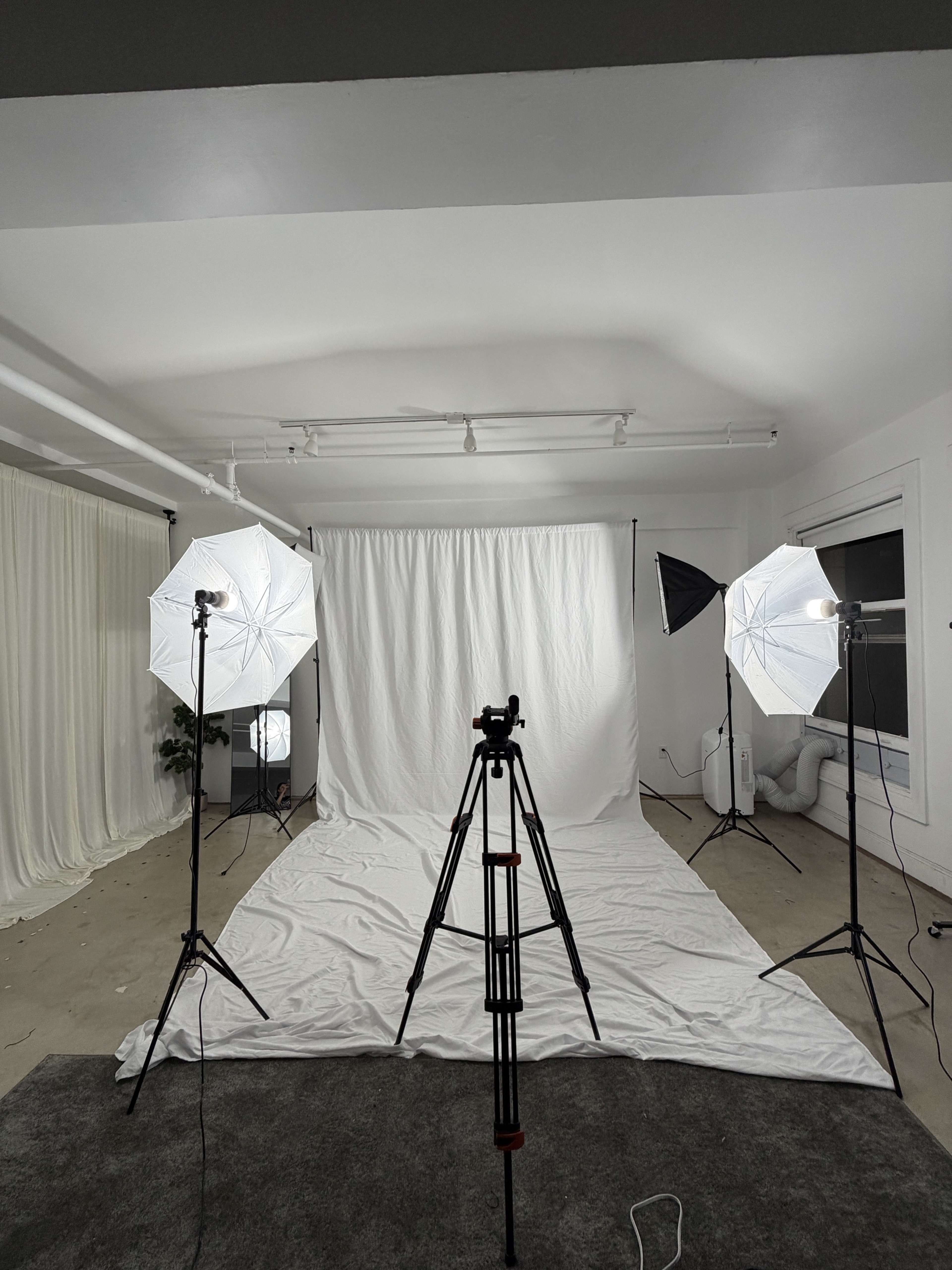The image shows a photography studio setup featuring a white backdrop, two large umbrella lights, and a tripod positioned on a textured floor.