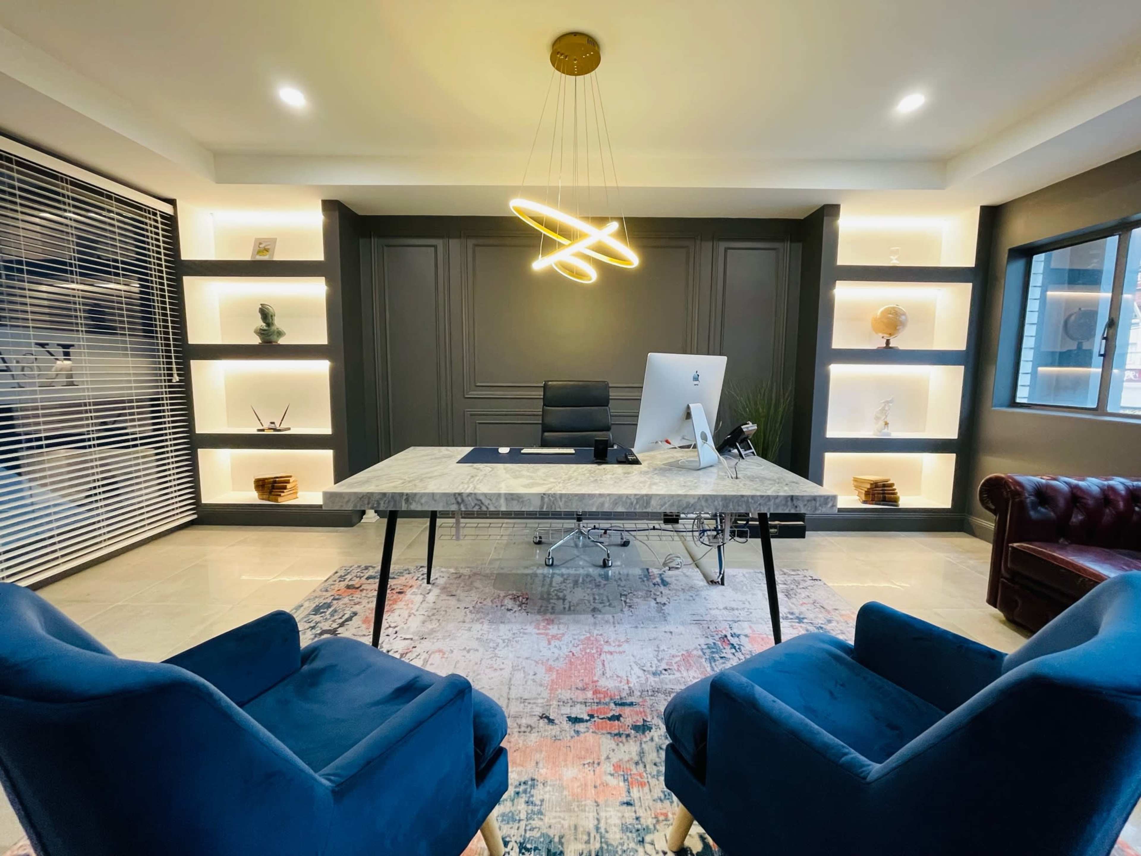 A modern office features a large marble desk with a computer, surrounded by two blue chairs, and illuminated by stylish overhead lighting.