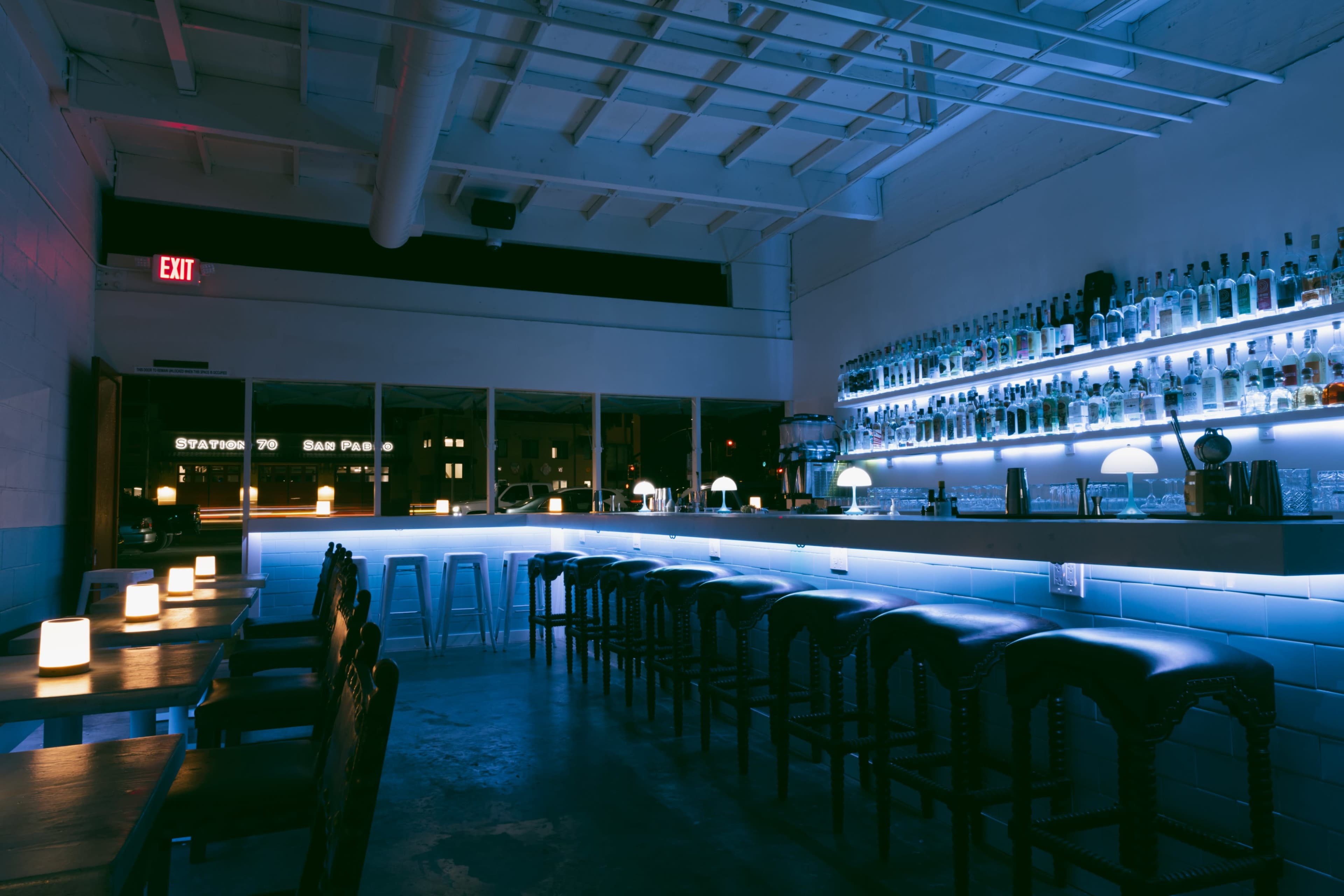 The image shows a modern bar interior featuring a long illuminated counter with stools, shelves lined with bottles, and a few tables with candles.
