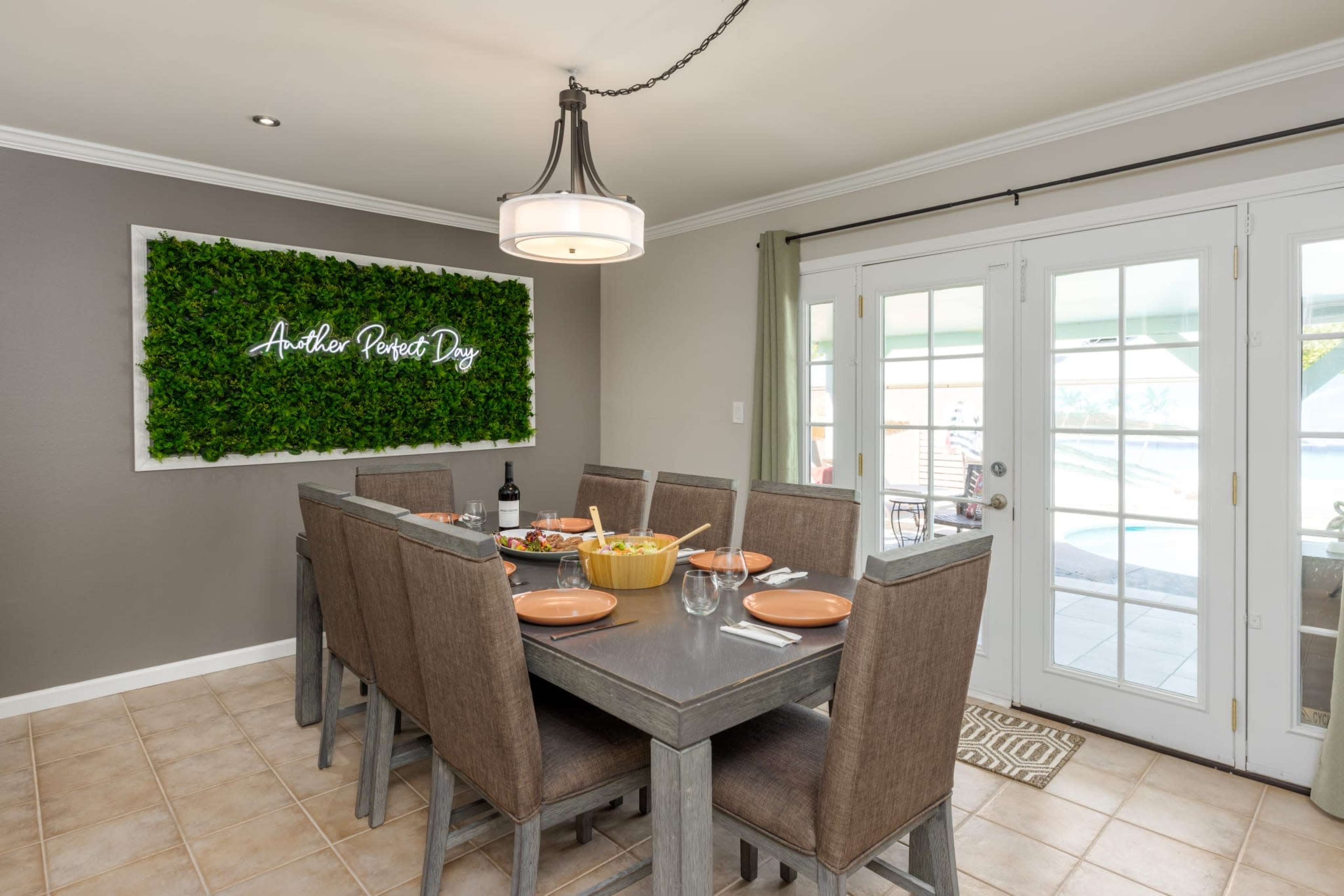 A dining area features a large table set for a meal, with a green wall decoration that says "Another Perfect Day" and doors leading outside.