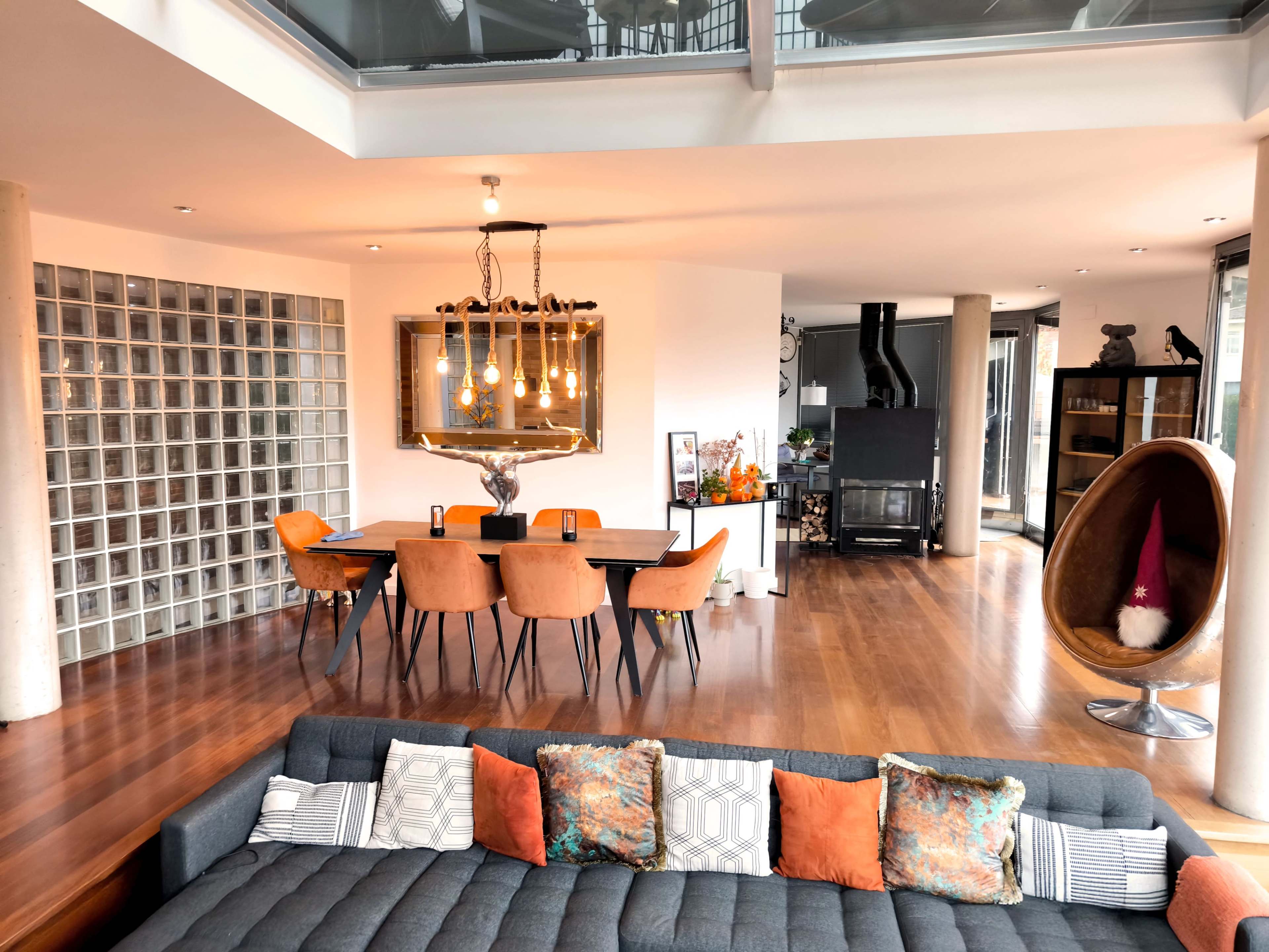 The image shows a modern living space with a gray sectional sofa in the foreground, a dining area with orange chairs and a chandelier in the center, and a fireplace visible in the background.
