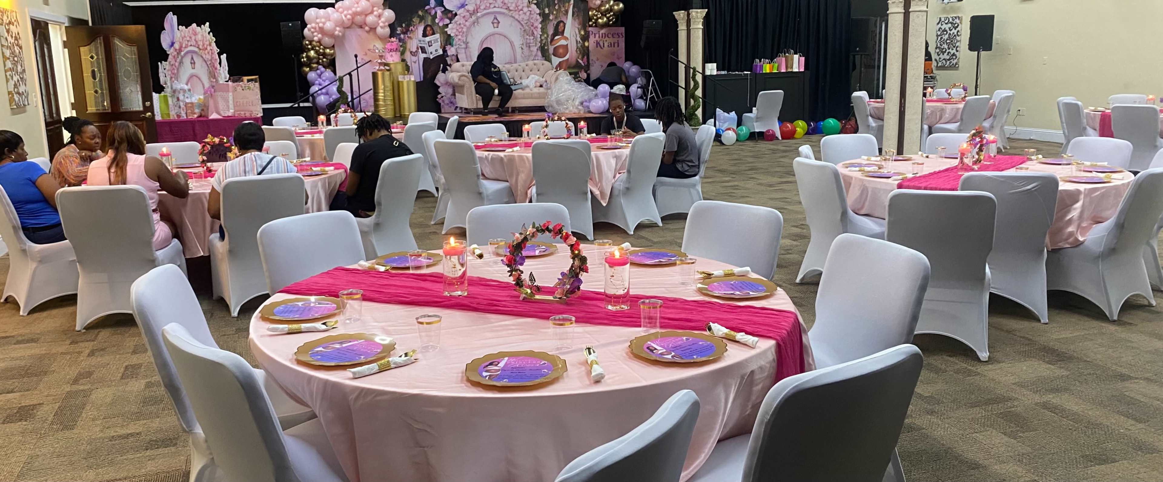 A banquet hall is set up with round tables covered in pink tablecloths, each adorned with centerpieces and plates, while decorations and balloons fill the background.