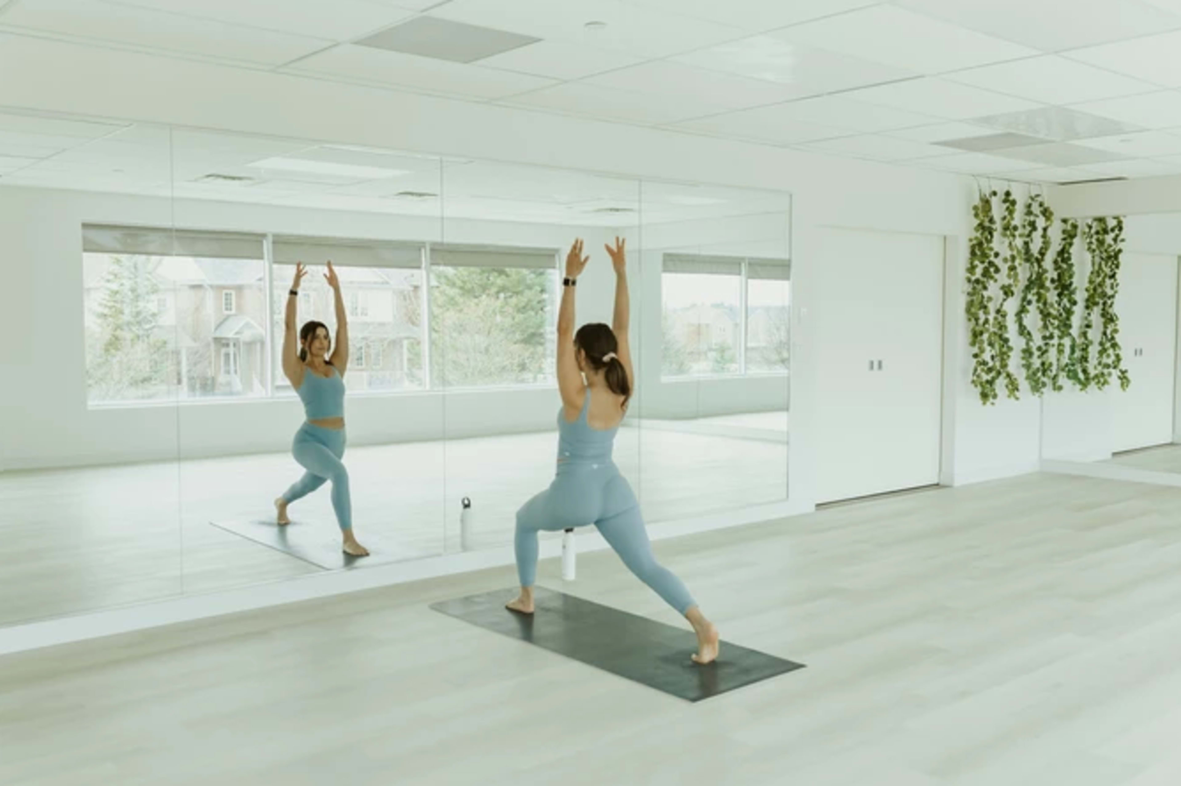 A person practices yoga in a mirrored studio with large windows and green plants on the wall.