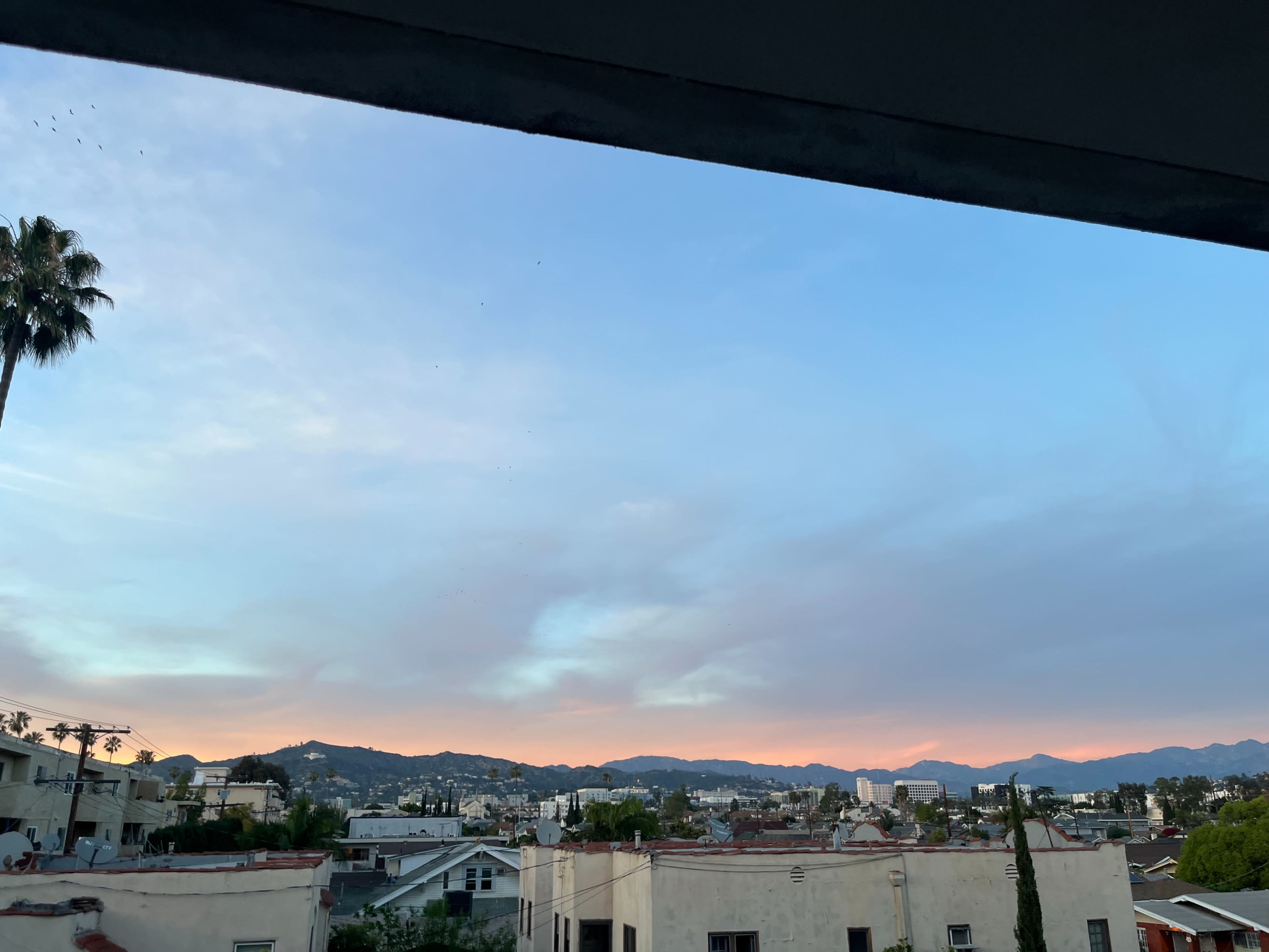 The image shows a view from a window, featuring a colorful sky at dusk above a cityscape with mountains in the background.