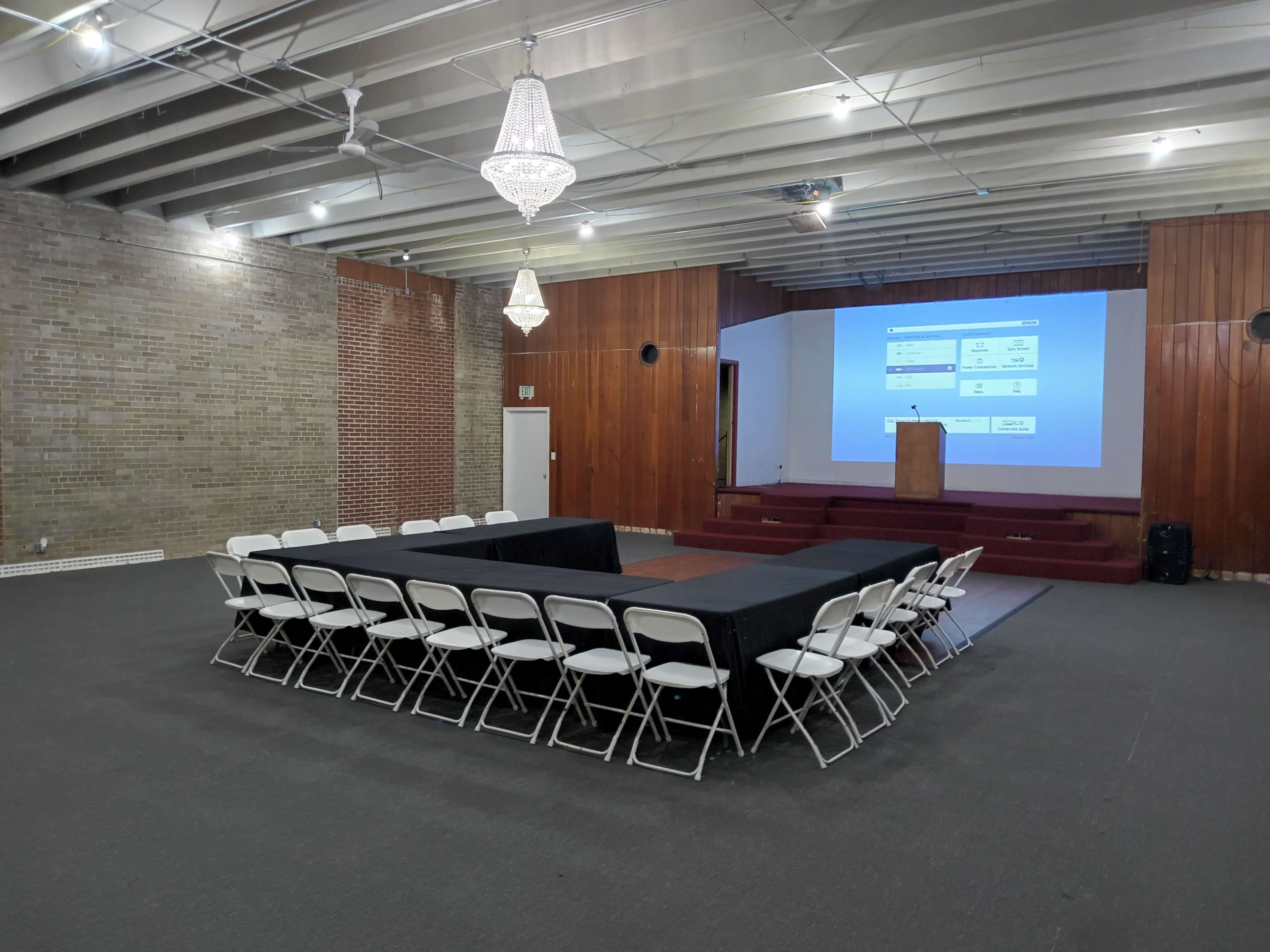 A conference room is set up with a rectangular arrangement of tables and chairs facing a presentation screen and podium.