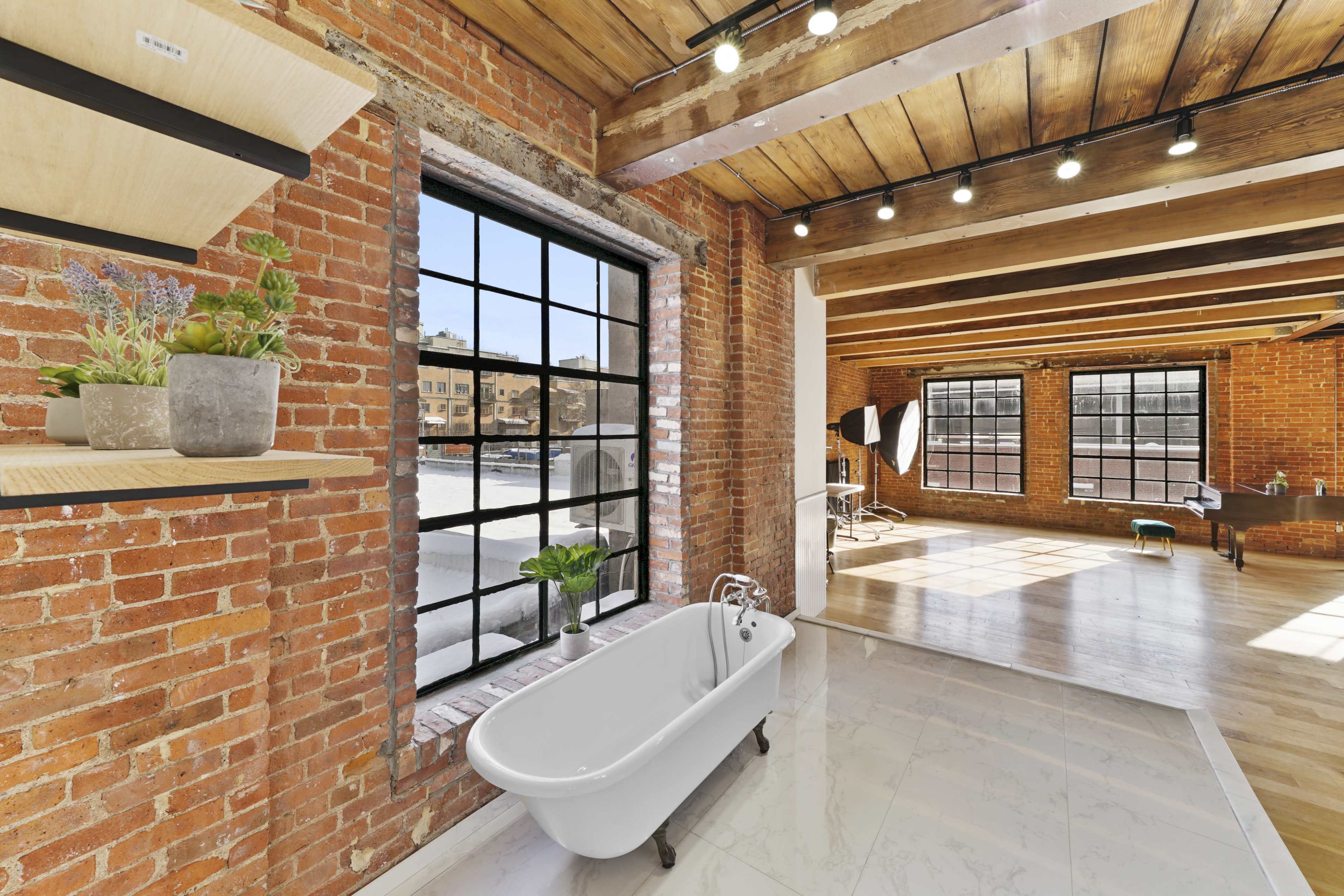 A spacious room with exposed brick walls, featuring a white freestanding bathtub and large windows that allow natural light to fill the space.