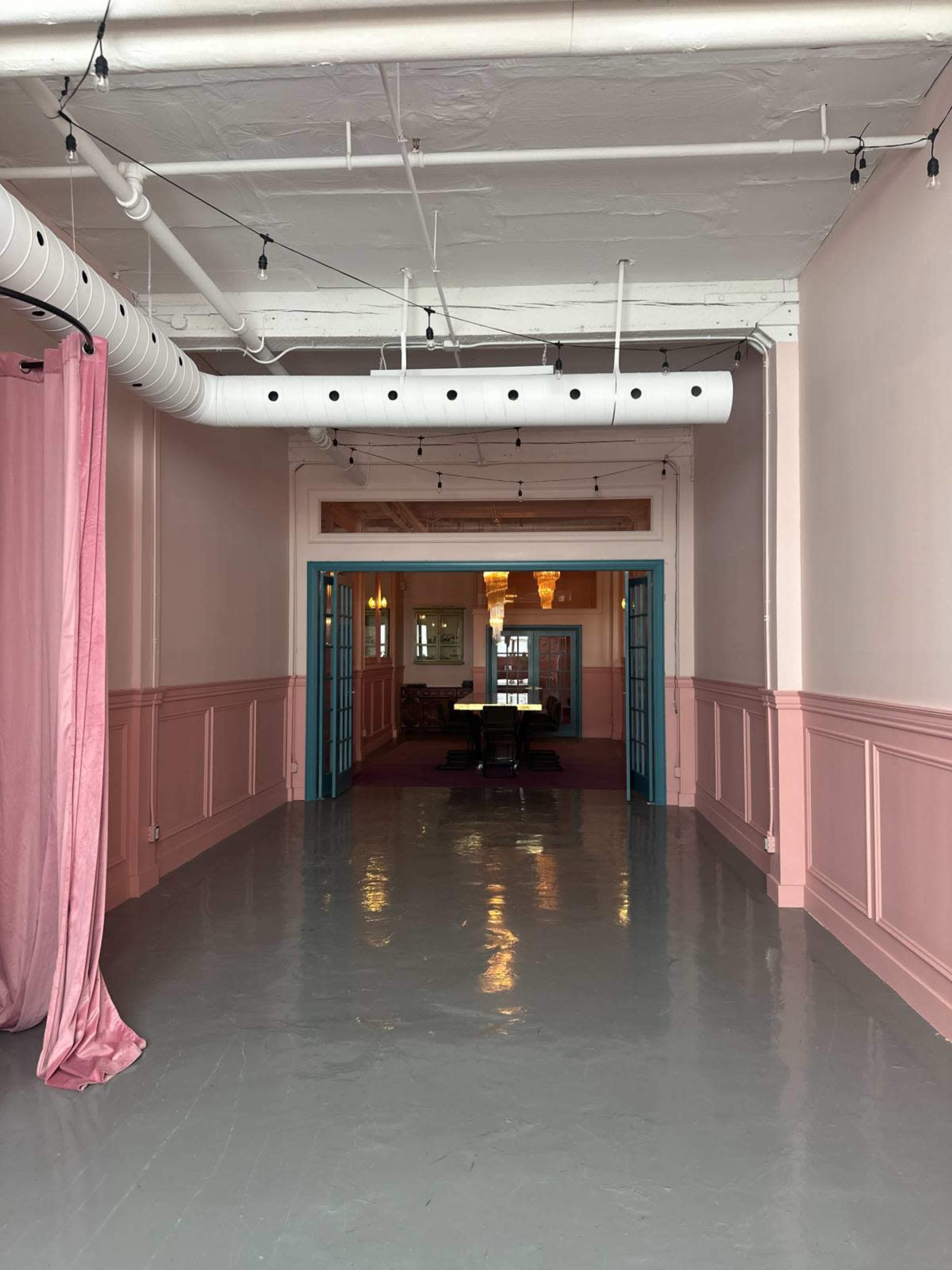 The image shows a wide corridor with pink walls and teal doors leading to a furnished room at the end, with decorative string lights overhead.