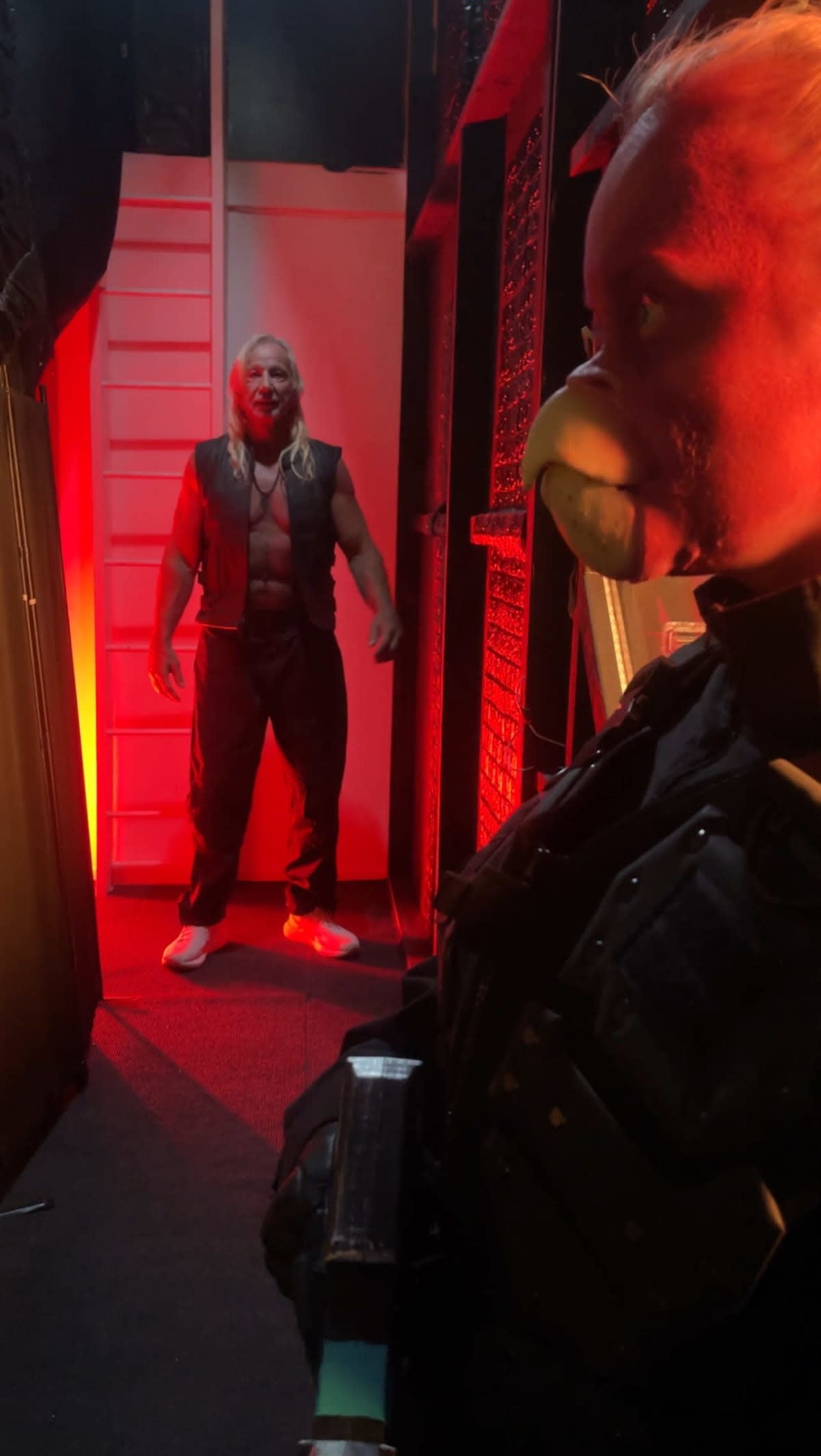 A muscular man with long hair and a vest stands menacingly in a dimly lit hallway, while a figure in tactical gear with a bird-like mask looks on.