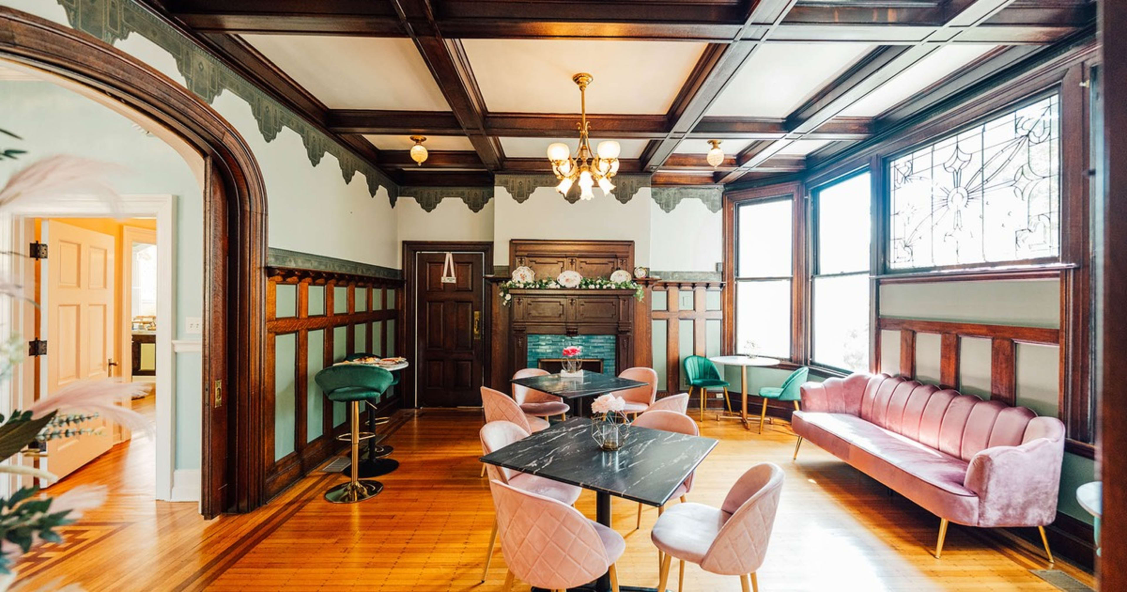 An elegantly furnished room with dark wooden paneling, a fireplace, and various seating arrangements, including pink and green chairs around a dark marble table.