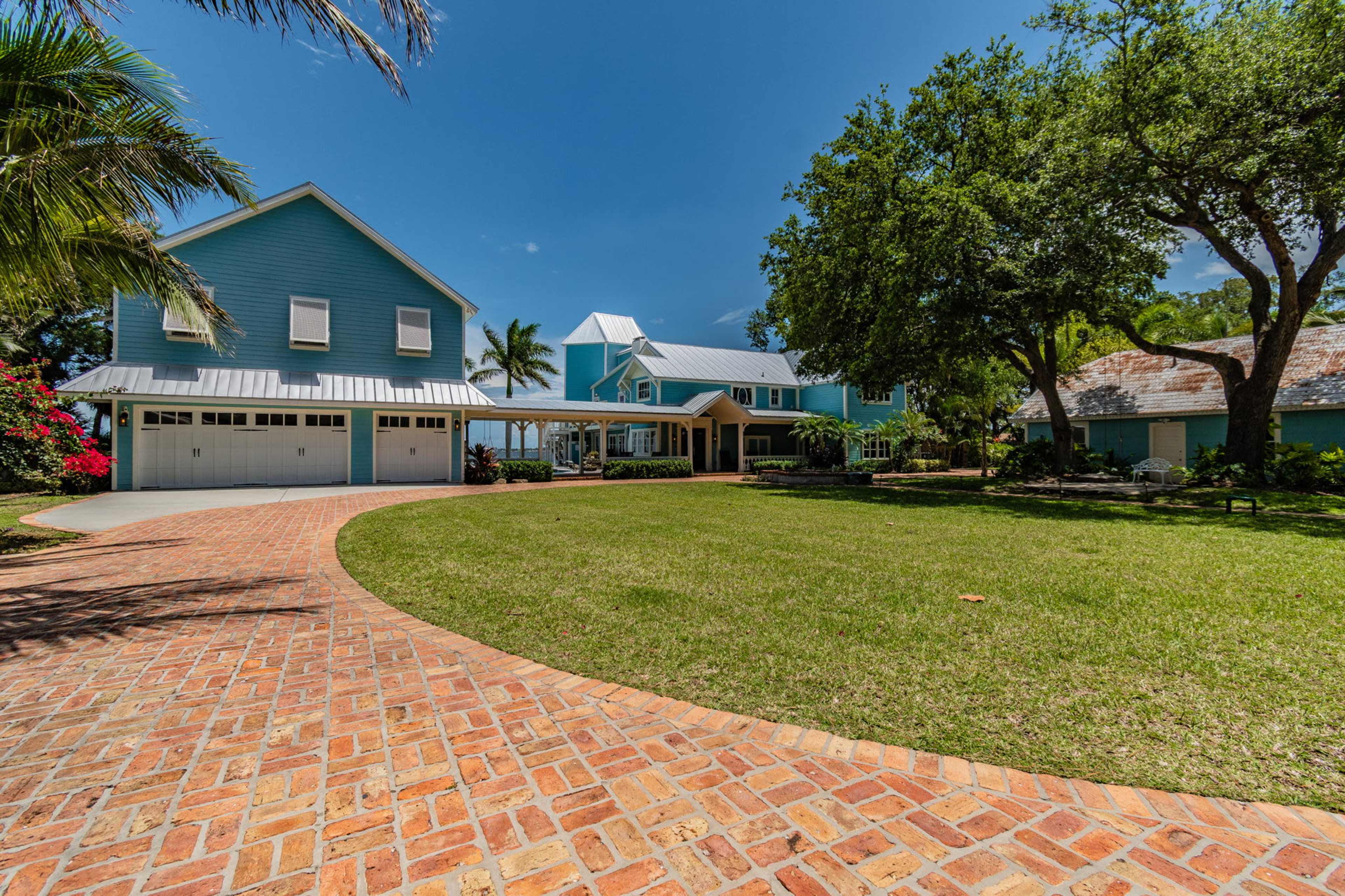 A large blue house with multiple gables, surrounded by a well-kept lawn and palm trees, with a brick pathway leading to the front entrance.