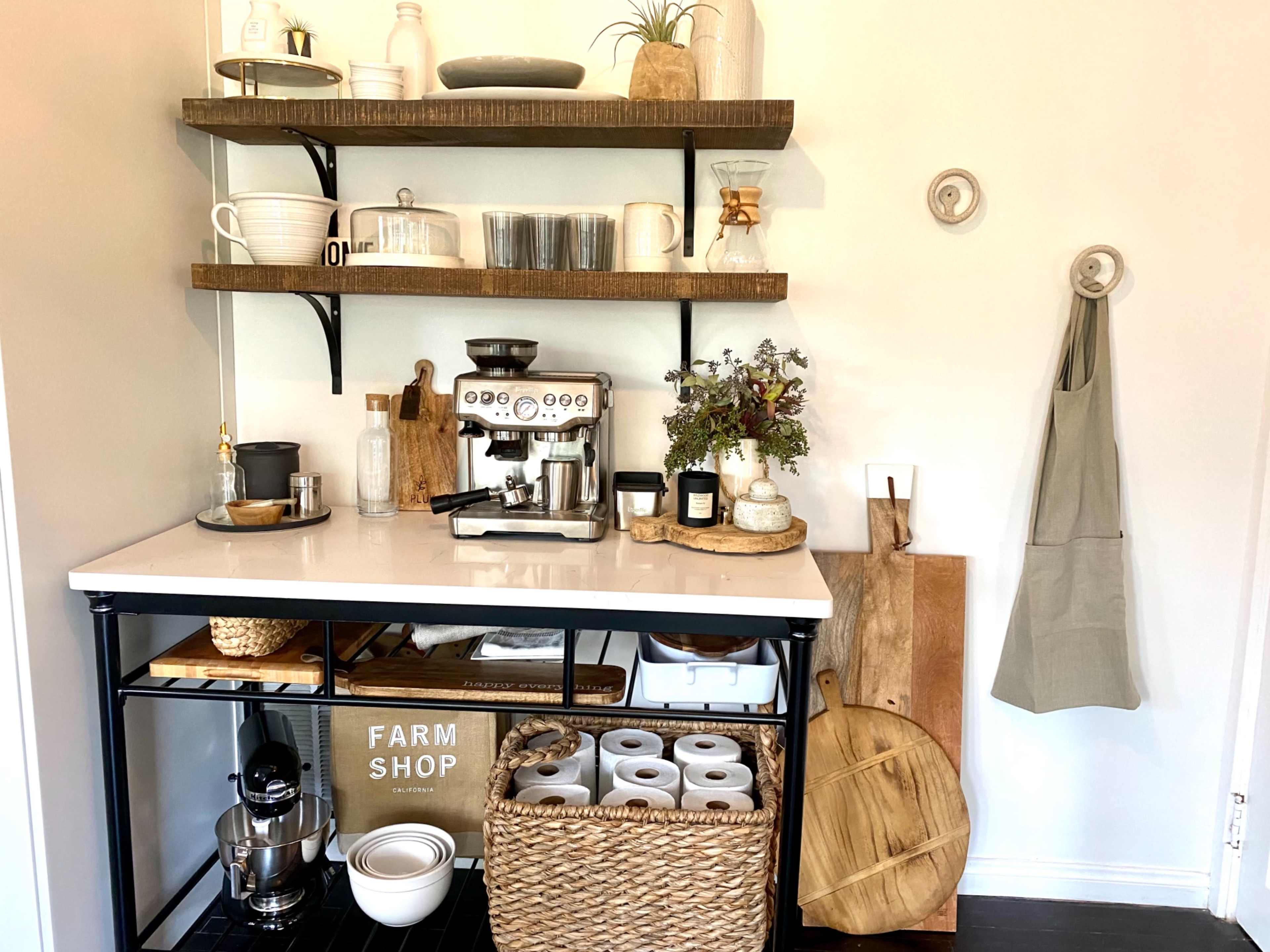 A coffee station with a machine on a black cart, surrounded by shelves holding dishes and decor, and a basket storing paper towels.