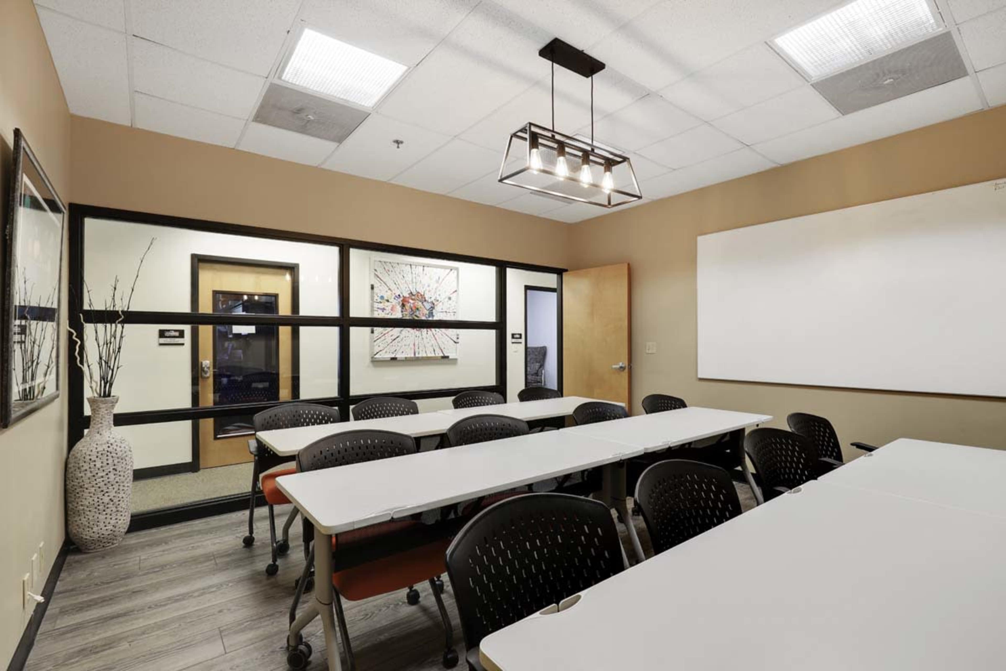 The image shows a modern conference room with several tables arranged in a U-shape, a large whiteboard, and a decorative vase in the corner.