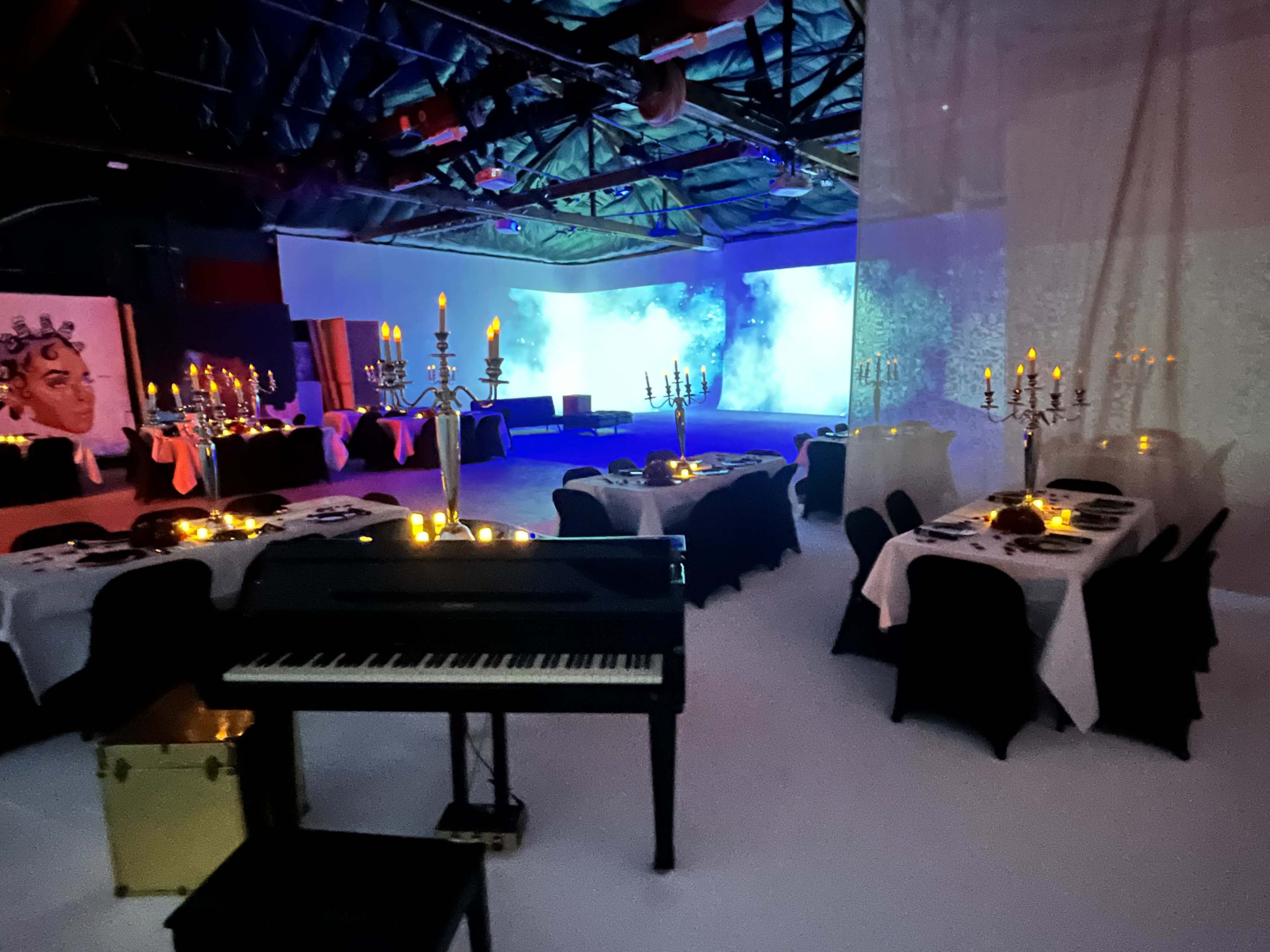 A banquet space features elegantly set tables with candles, a grand piano, and a vibrant light display on the wall.