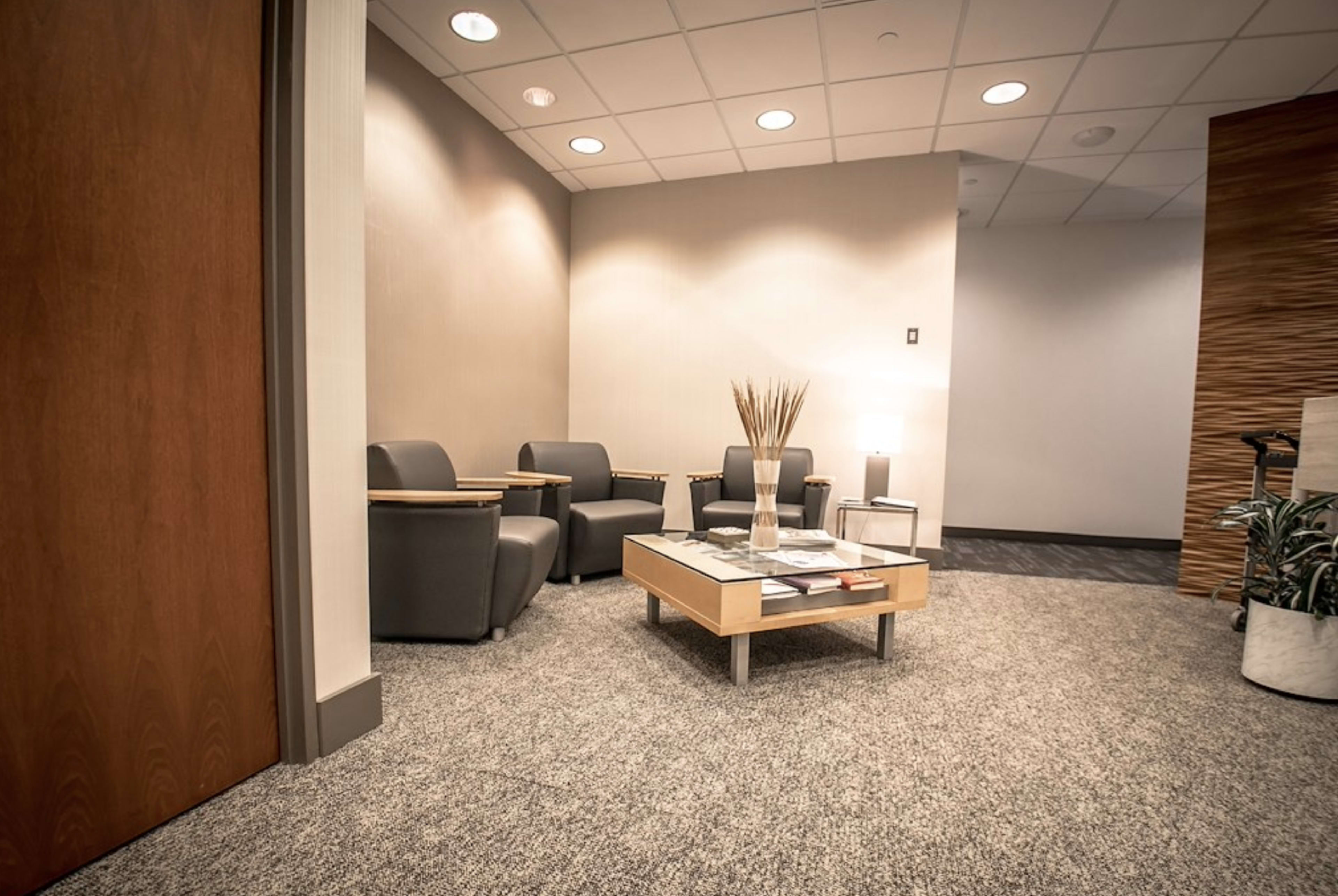 The image shows a modern waiting area with two gray chairs, a wooden table, and a decorative plant in a corner.