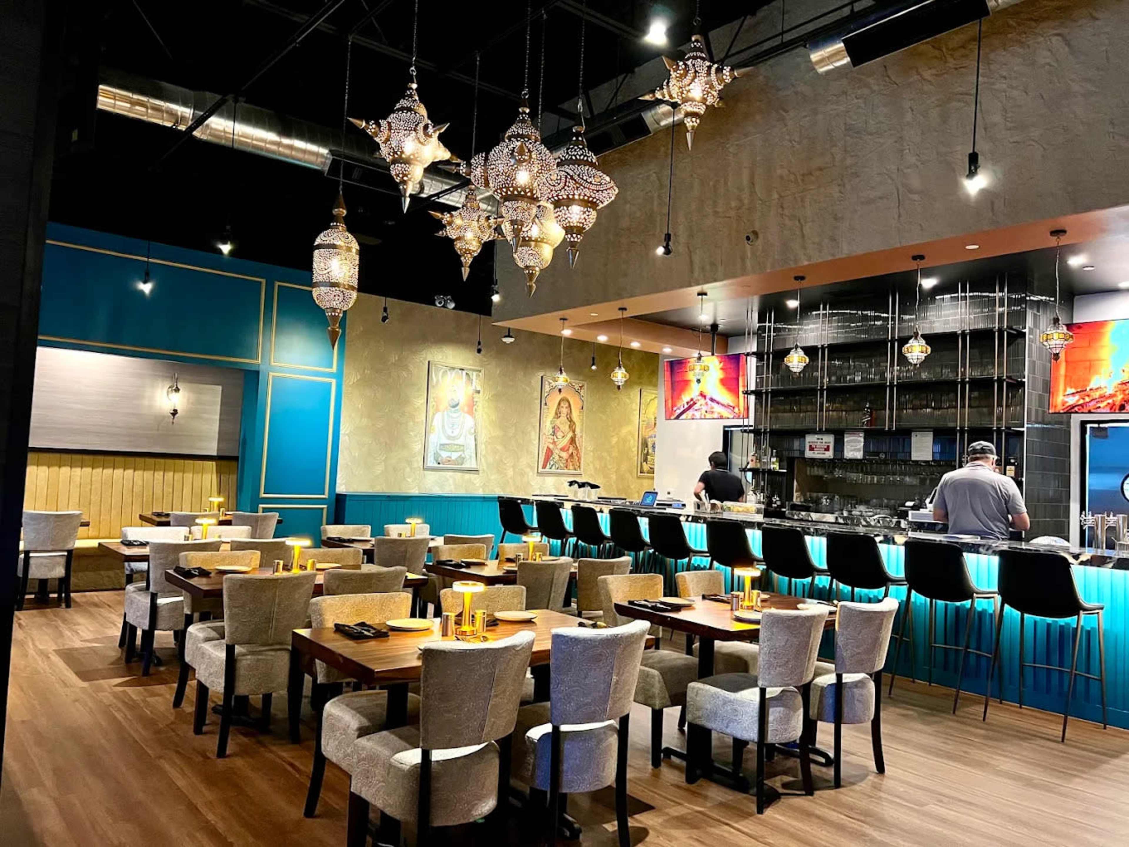 The image depicts a modern restaurant interior with elegant lighting, featuring a bar area, tables arranged in a dining space, and chefs preparing food in the background.