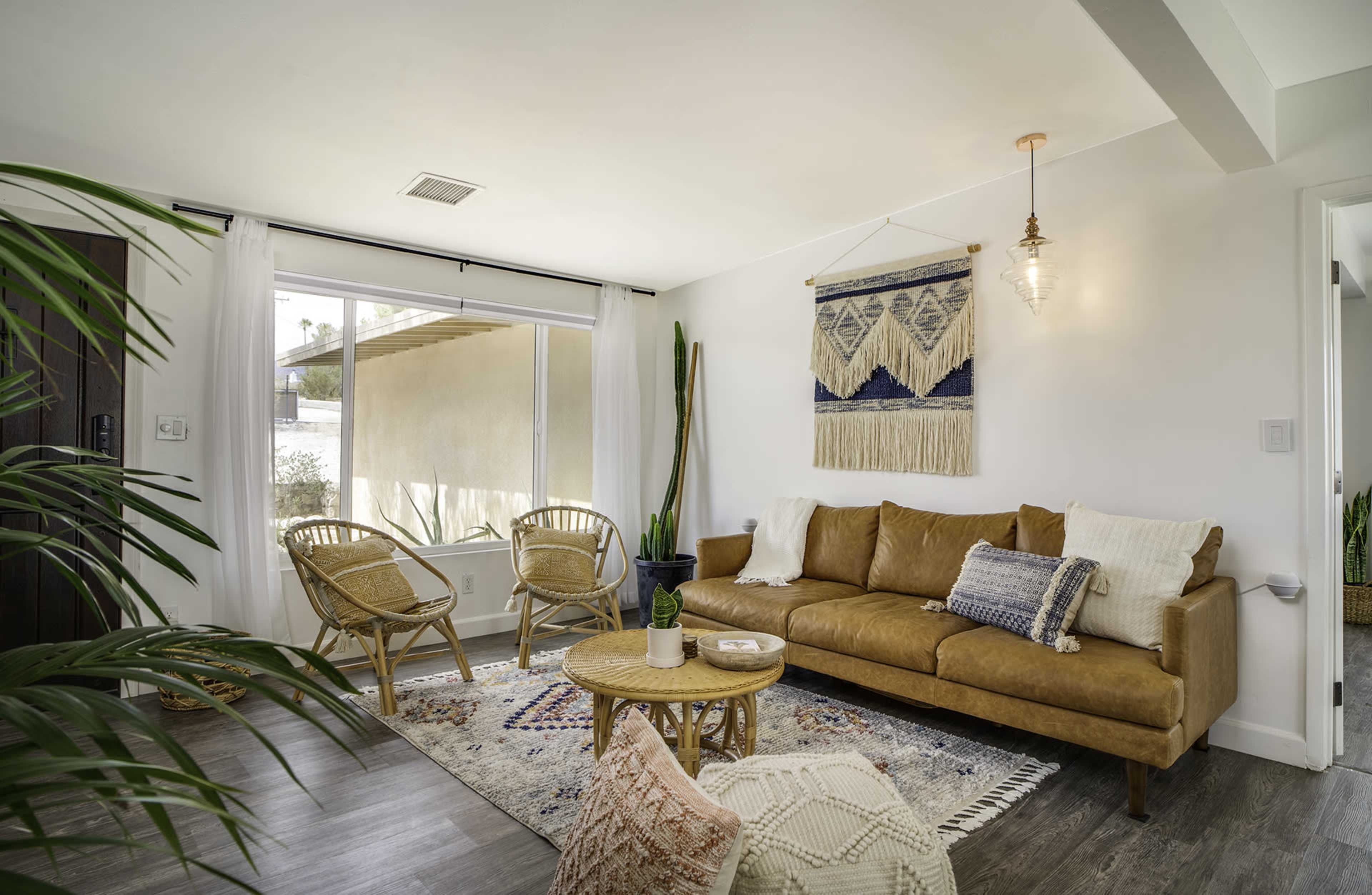 A cozy living room features a brown leather sofa, a round wooden coffee table, and rattan chairs, with large windows allowing natural light and greenery in the decor.