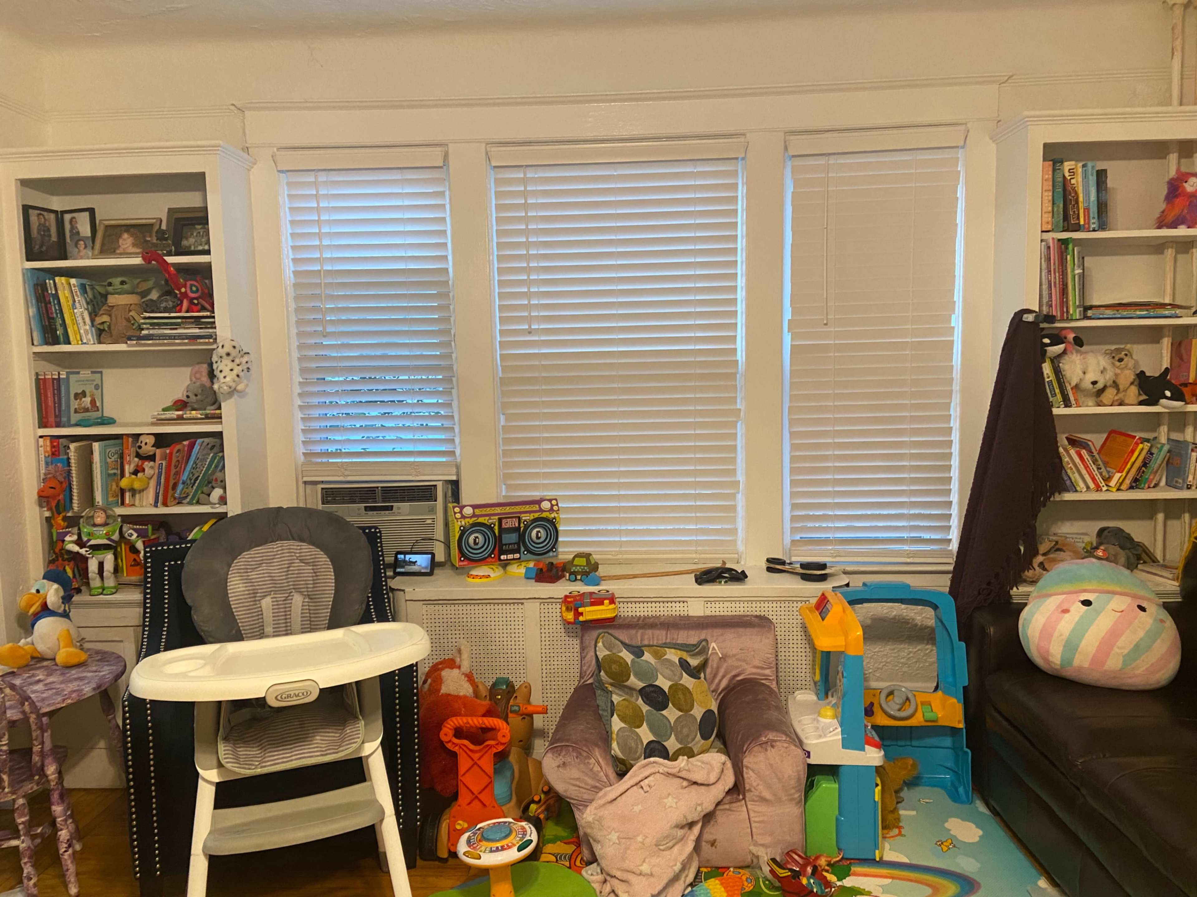 The image shows a cluttered living room with toys scattered across the floor and furniture, including a high chair and a pink armchair.