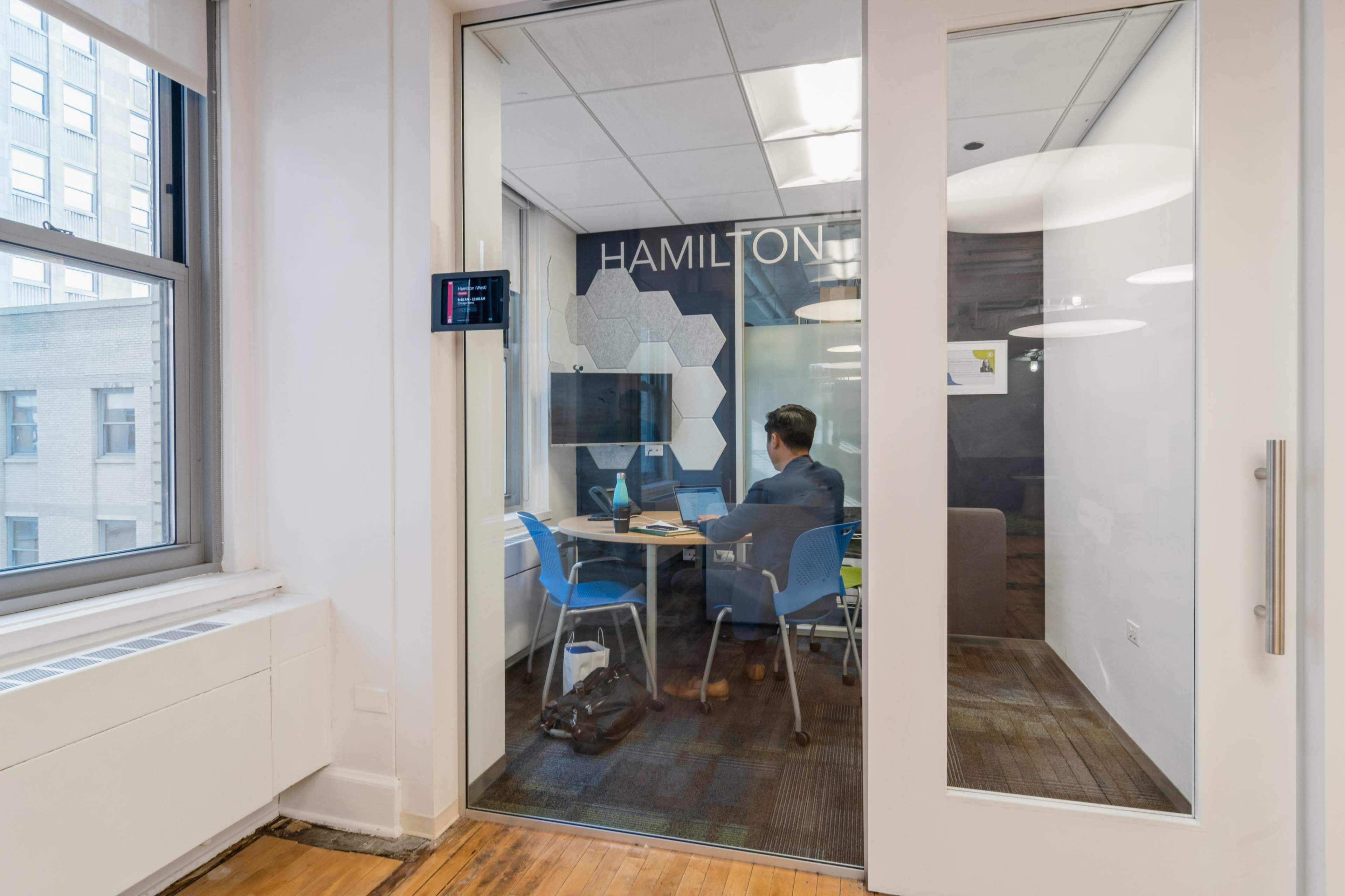 A person is working at a desk inside a glass meeting room labeled "HAMILTON" with blue chairs and modern decor.
