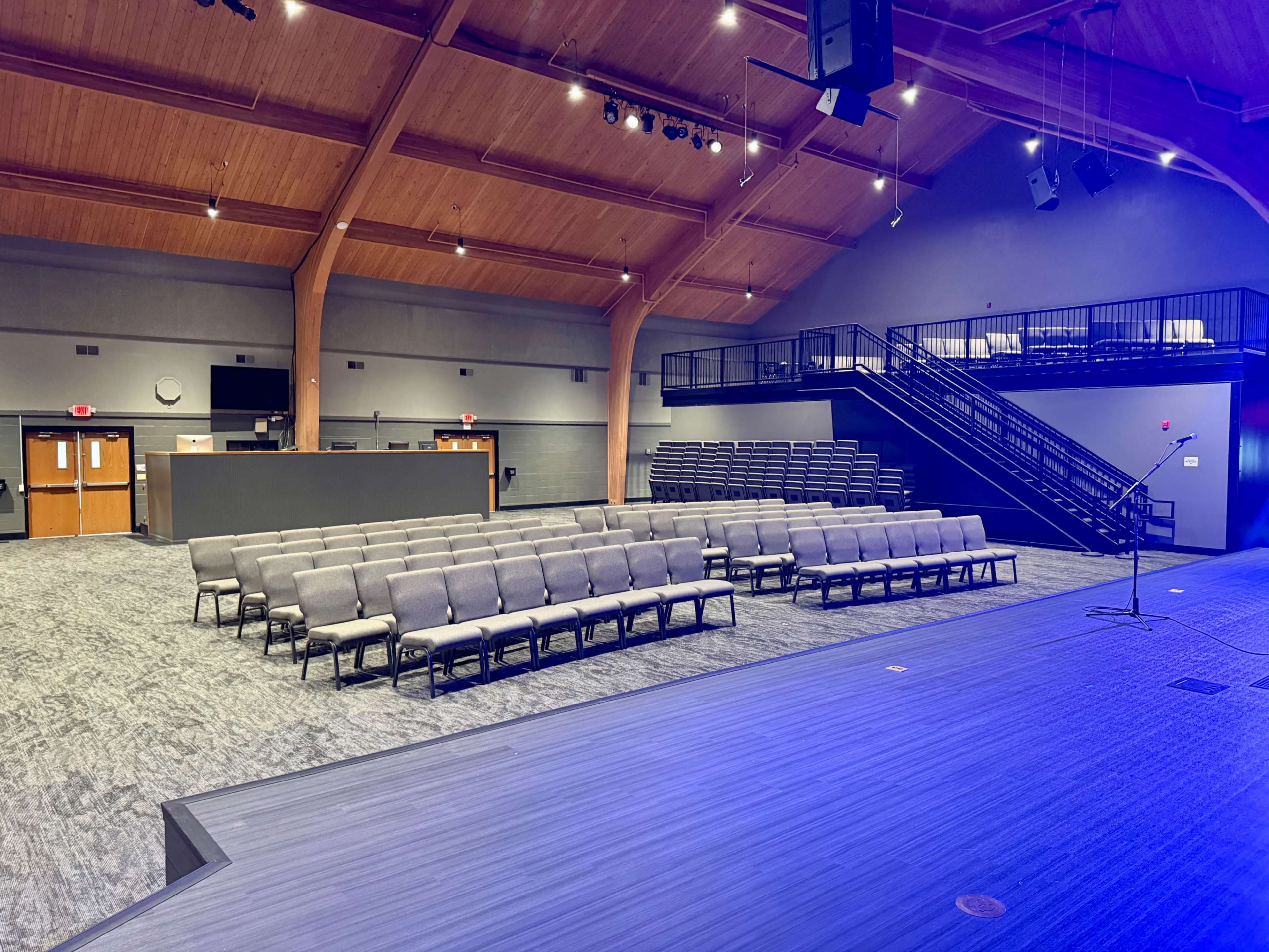 The image shows a spacious indoor venue with a stage, seating arranged in rows, and a balcony in the background.