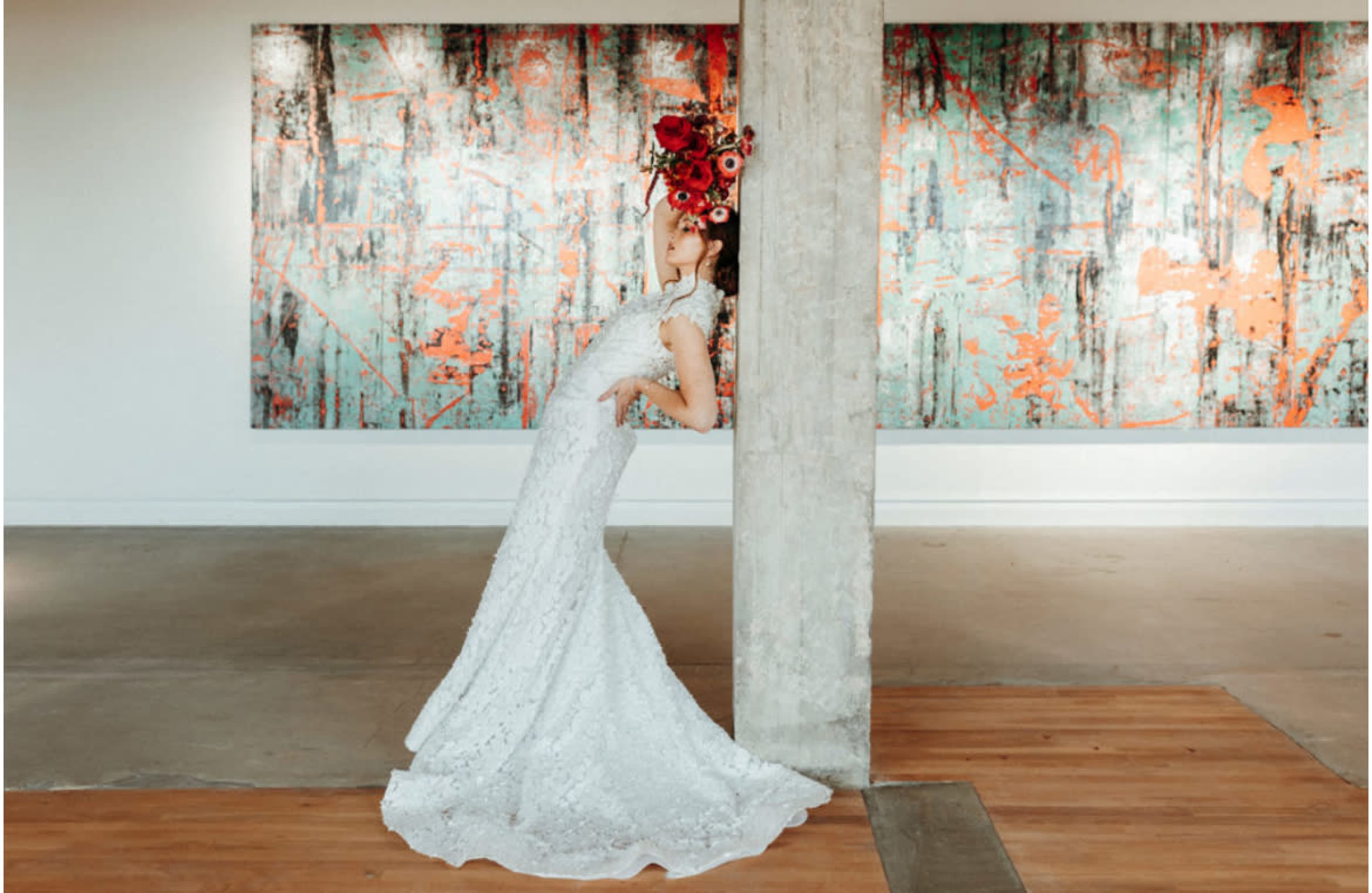 A woman in a white lace gown leans against a concrete pillar while adorned with a bouquet of flowers, set against colorful abstract art on the walls.