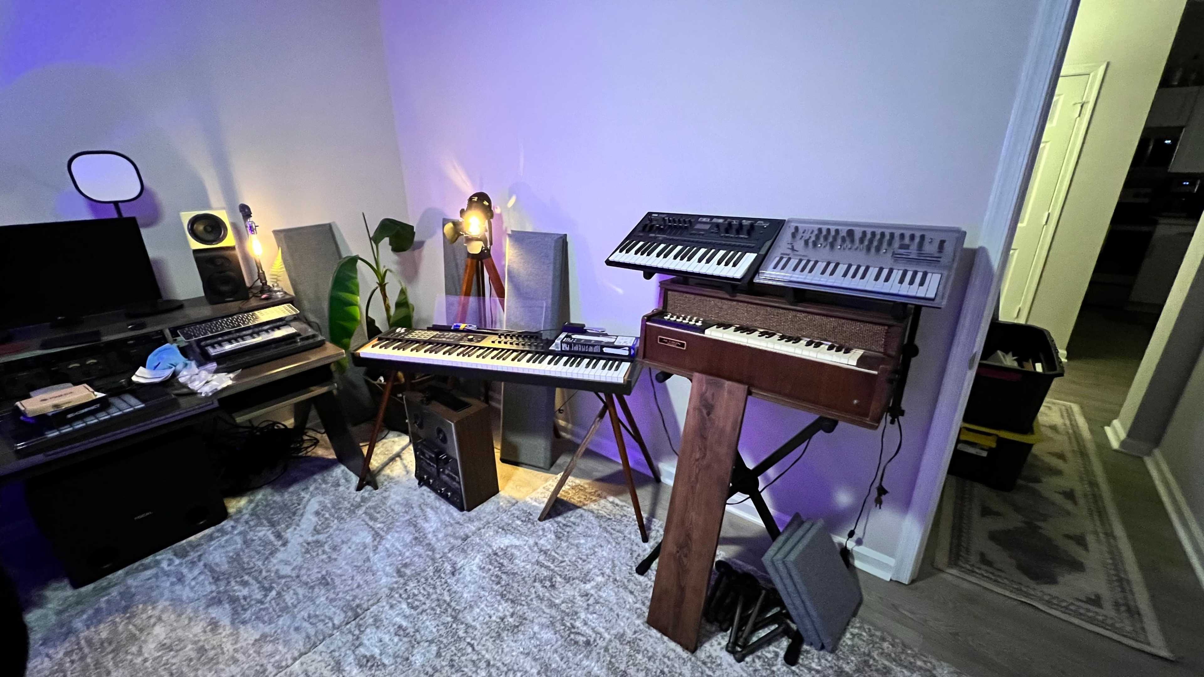 A music production corner with various synthesizers and keyboards arranged against a wall, illuminated by soft lighting.
