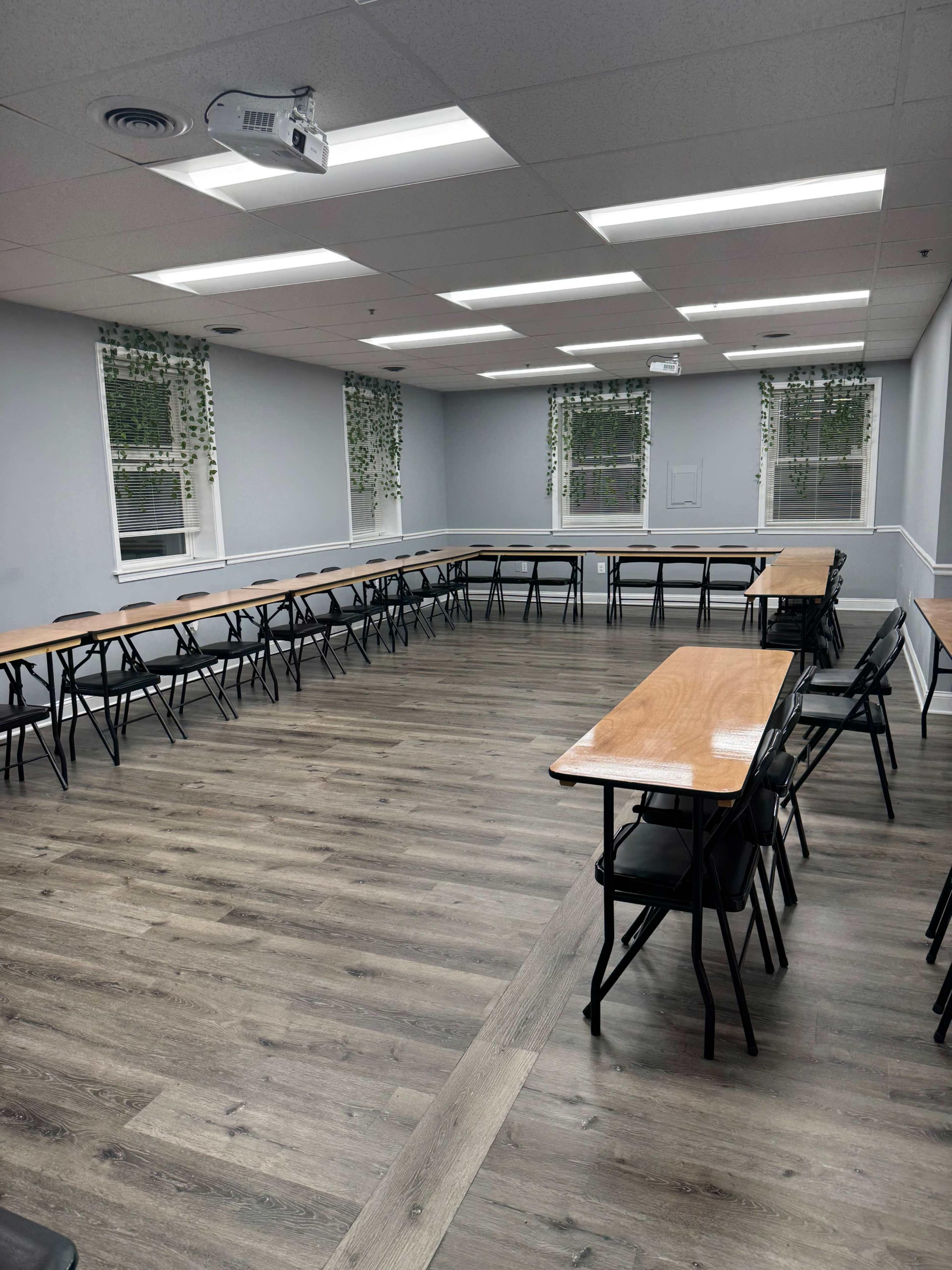 The image shows a classroom with multiple rectangular tables arranged in a U-shape, surrounded by black folding chairs, and featuring large windows adorned with greenery.