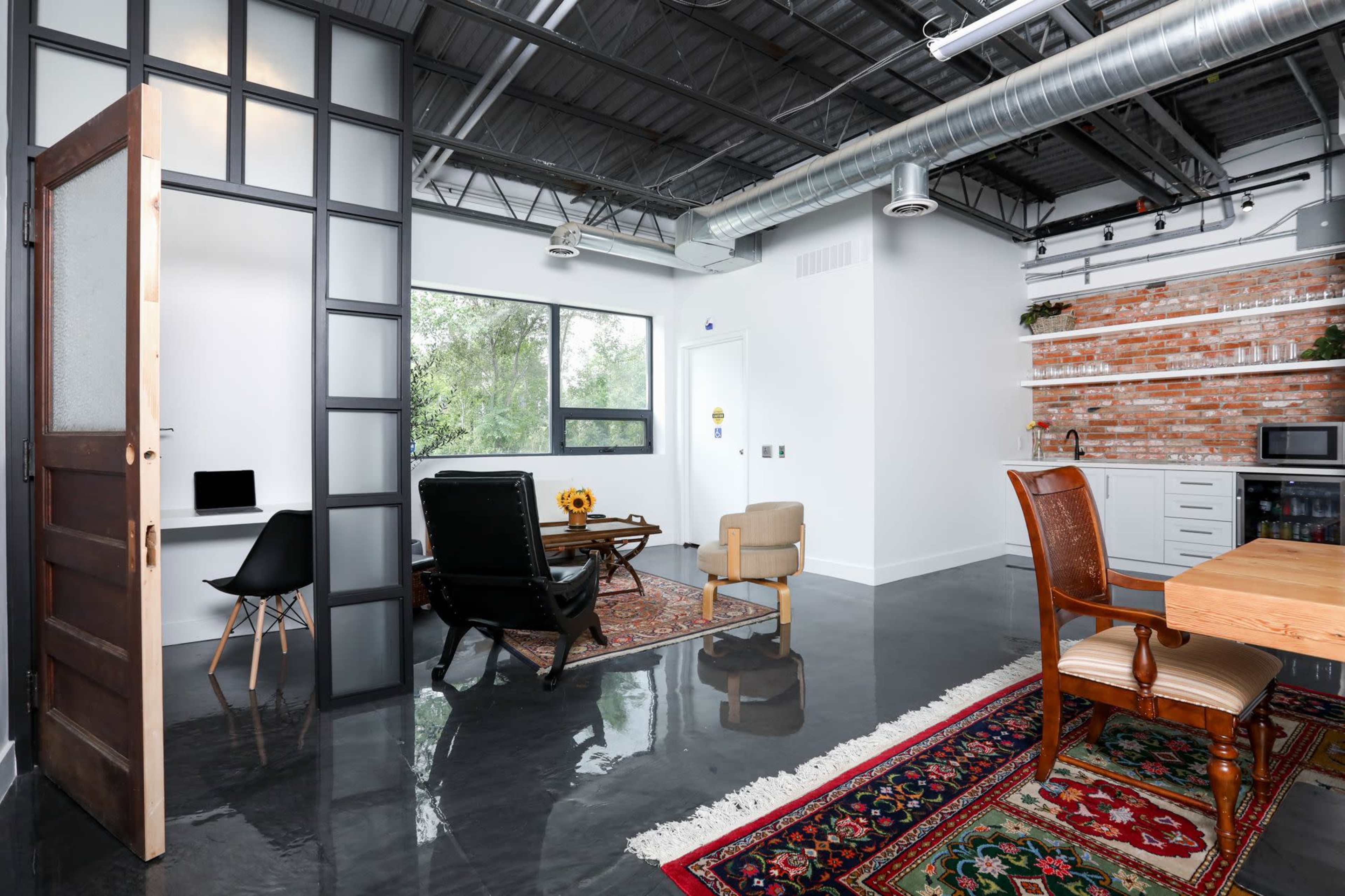 A modern office space with a wooden door, stylish furniture, and a kitchen area featuring brick accents.