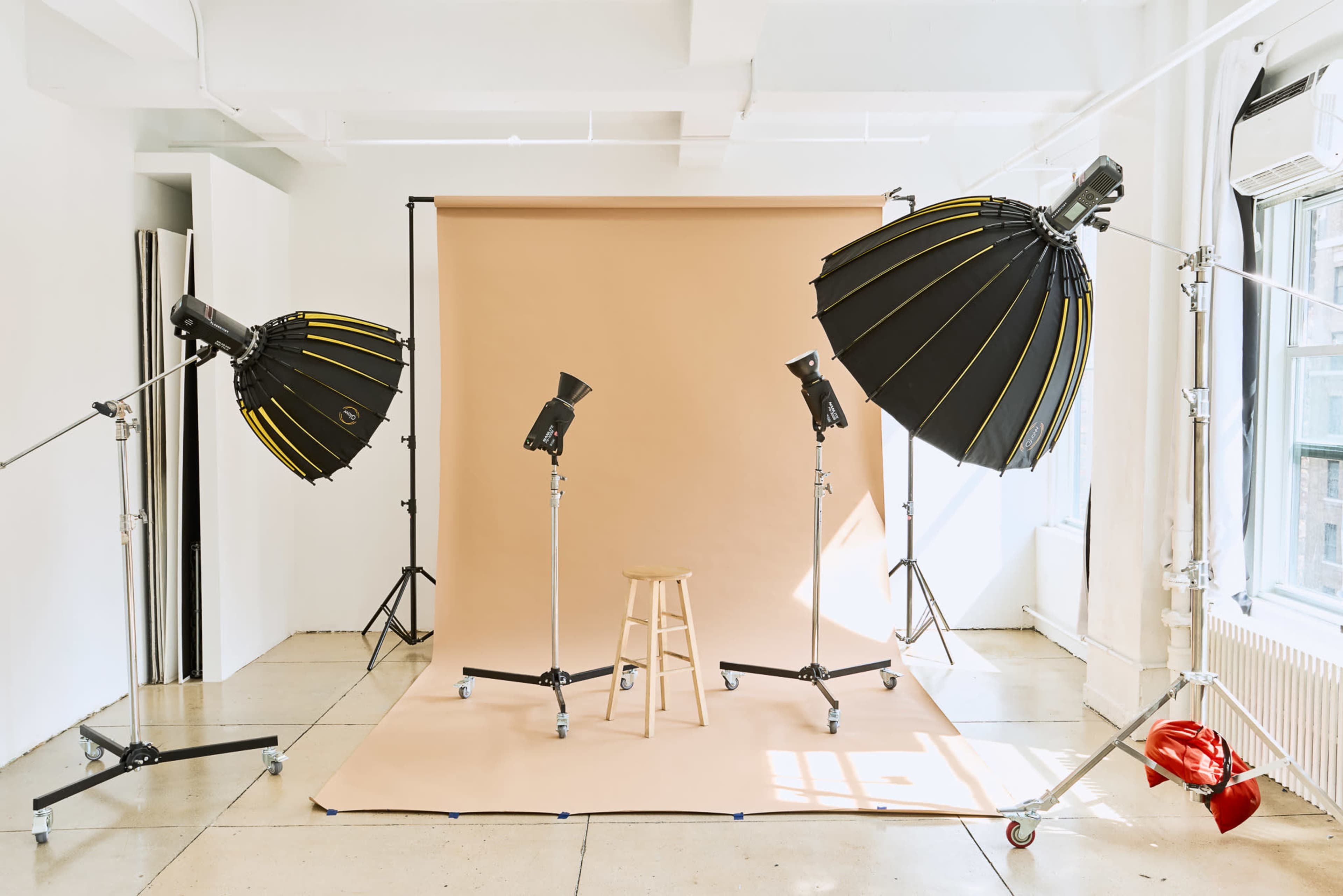 A photography studio features two large softbox lights and a stool set in front of a plain beige backdrop.
