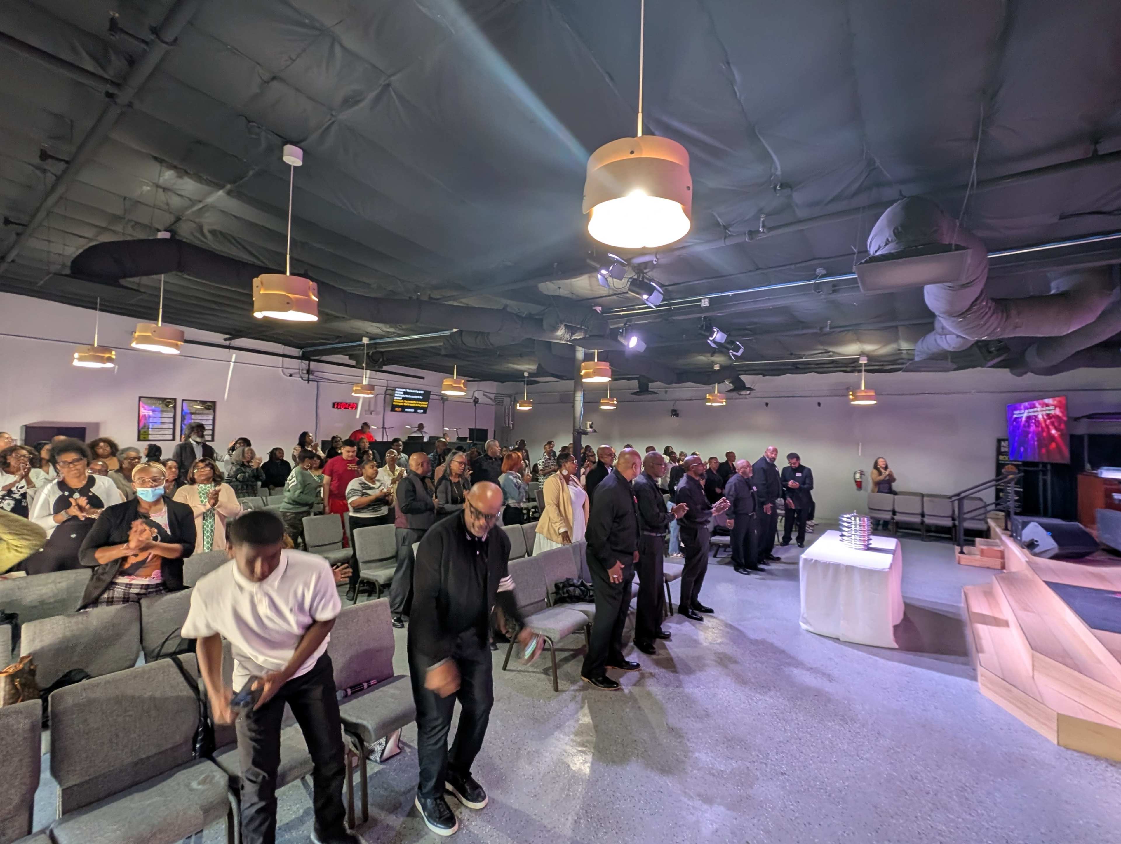A large group of people stands in a modern indoor space, with rows of chairs and a stage visible in the background.