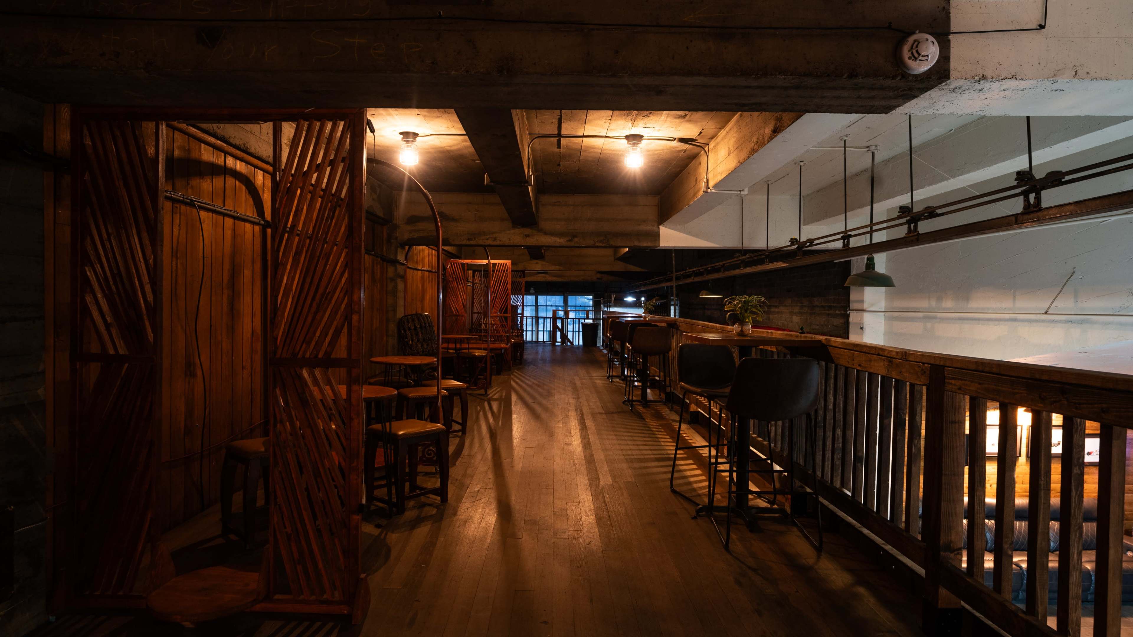 The image shows a dimly lit bar area with wooden decor, including tall stools and a railing along a long corridor.