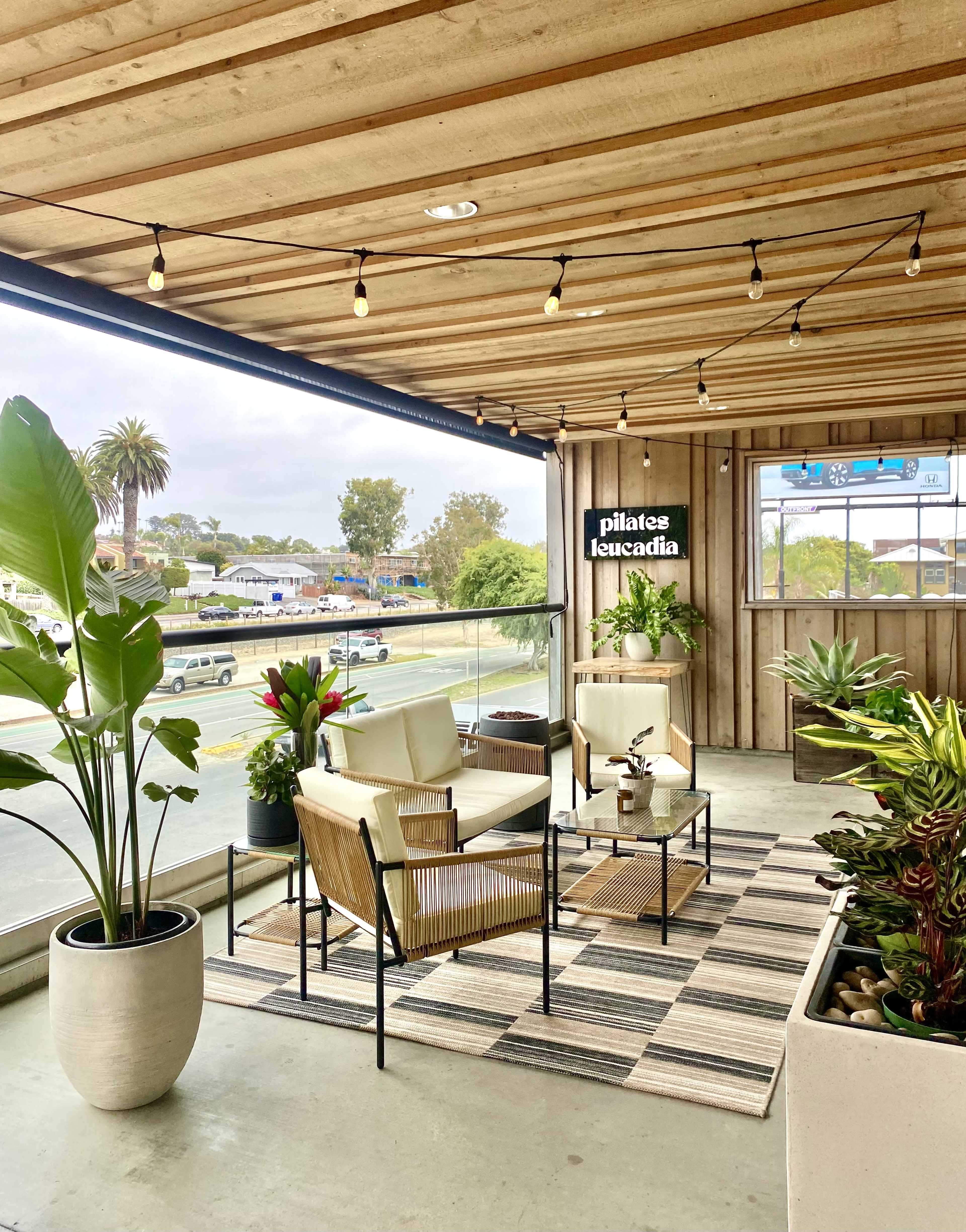The image shows a modern, well-decorated outdoor seating area with potted plants, a rug, and a sign for "Pilates Leucadia" against a wooden wall.