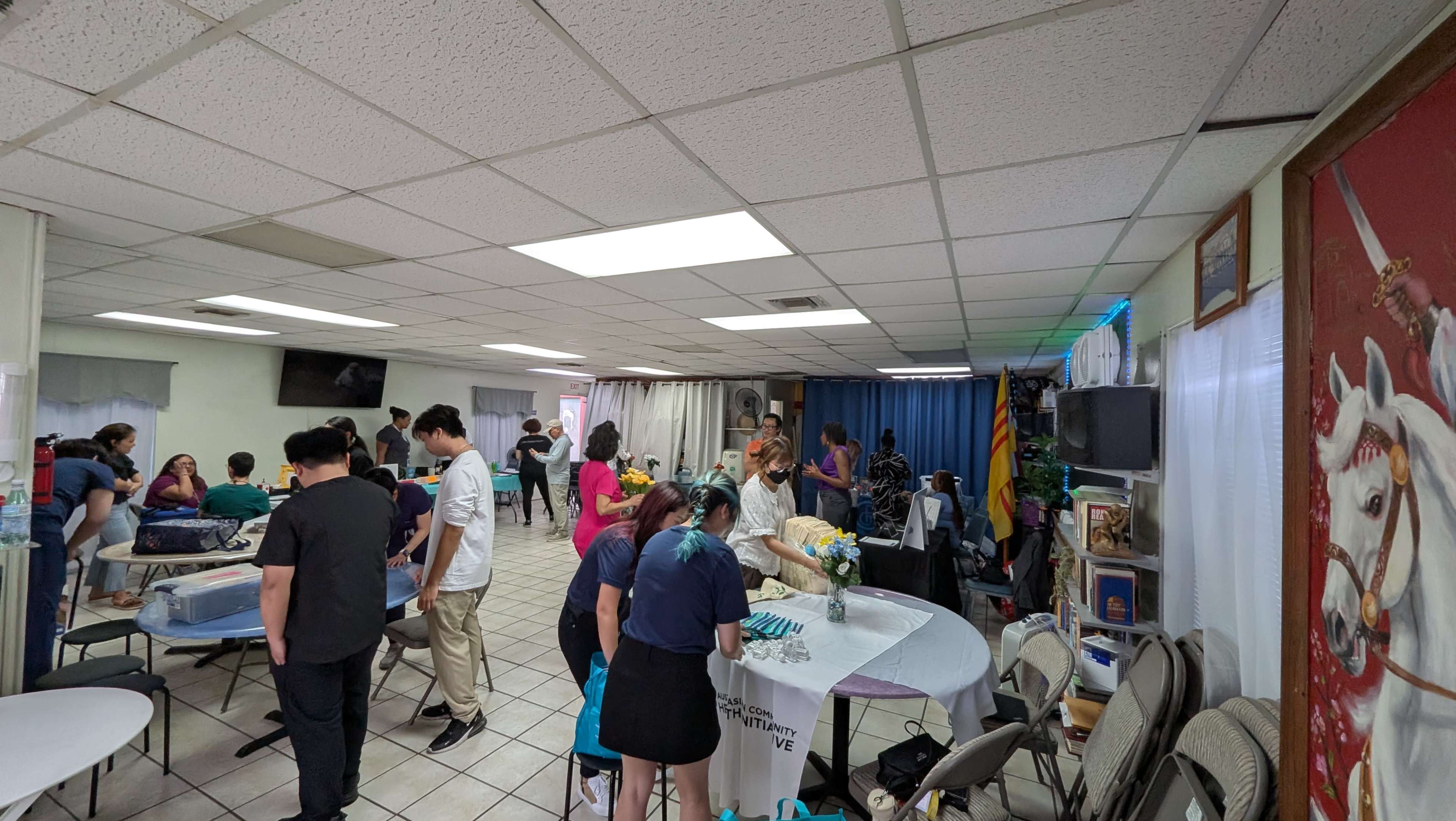 A large group of people is gathered inside a bright, spacious room, engaging in various activities around tables while decorations adorn the walls.