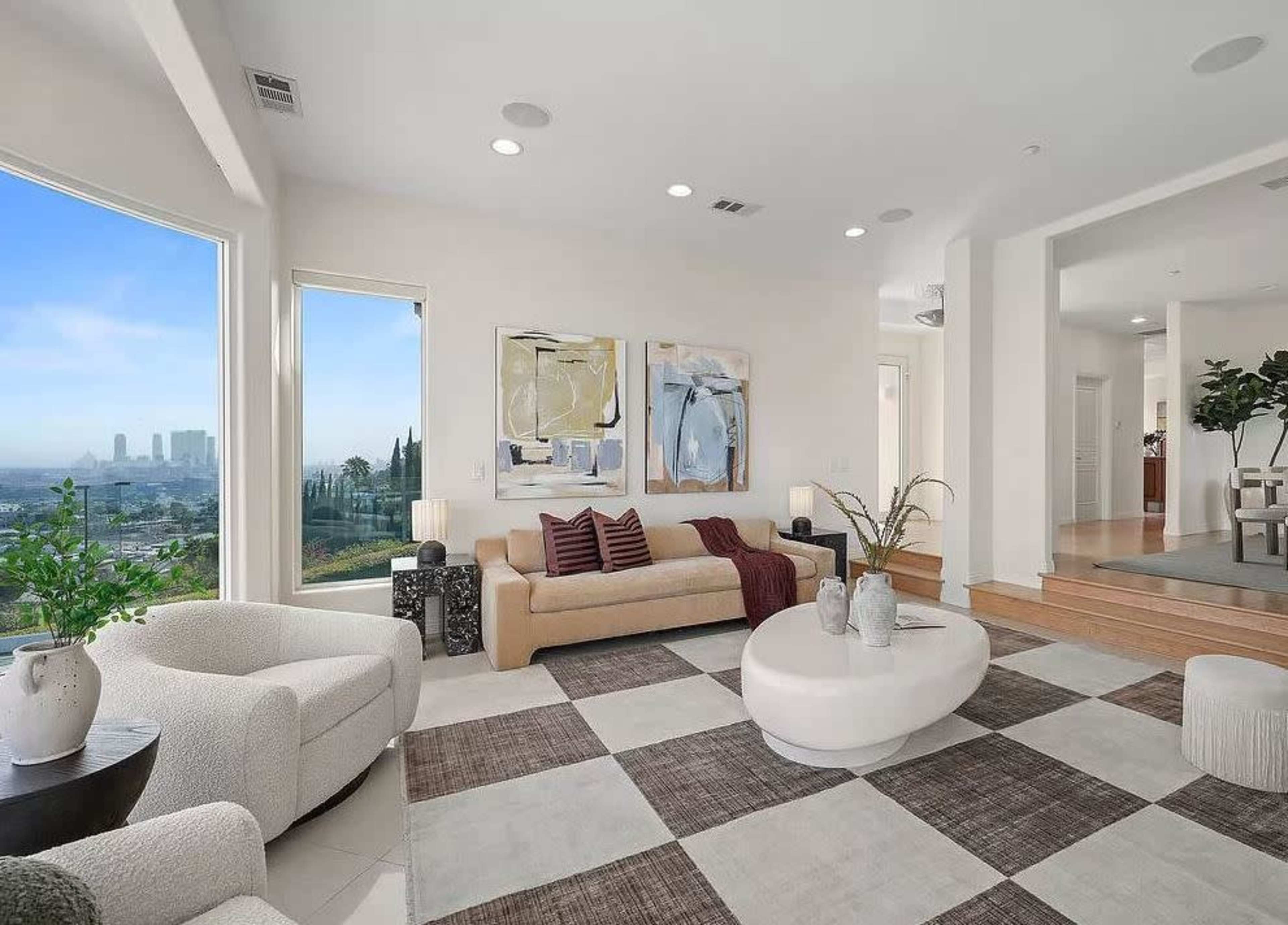 The image depicts a modern living room with large windows showcasing a city skyline, featuring a beige sofa, two light armchairs, and a circular coffee table on a patterned rug.