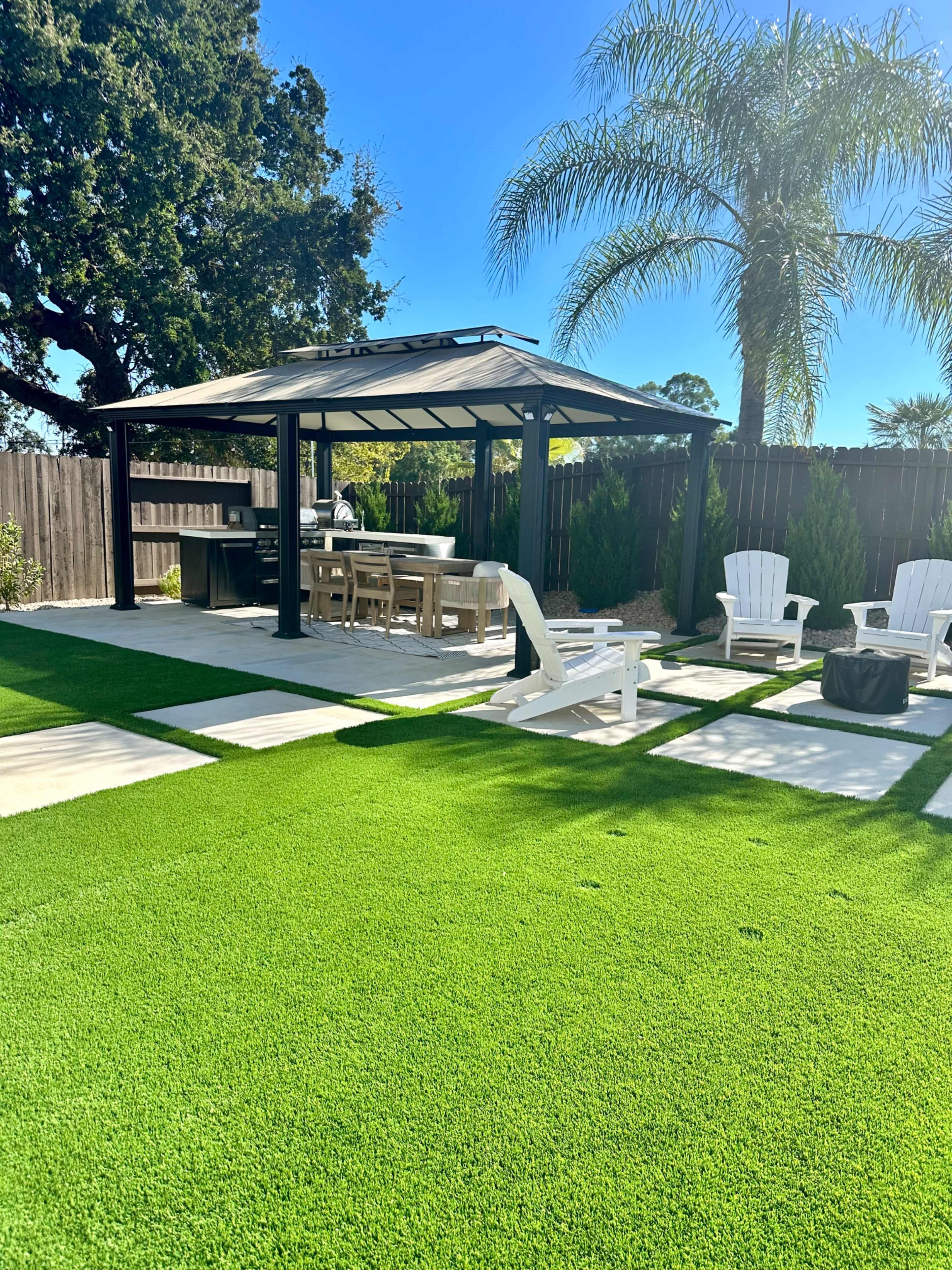 The image shows a well-maintained backyard featuring a gazebo with dining furniture, an outdoor kitchen area, and manicured artificial grass bordered by palm trees.