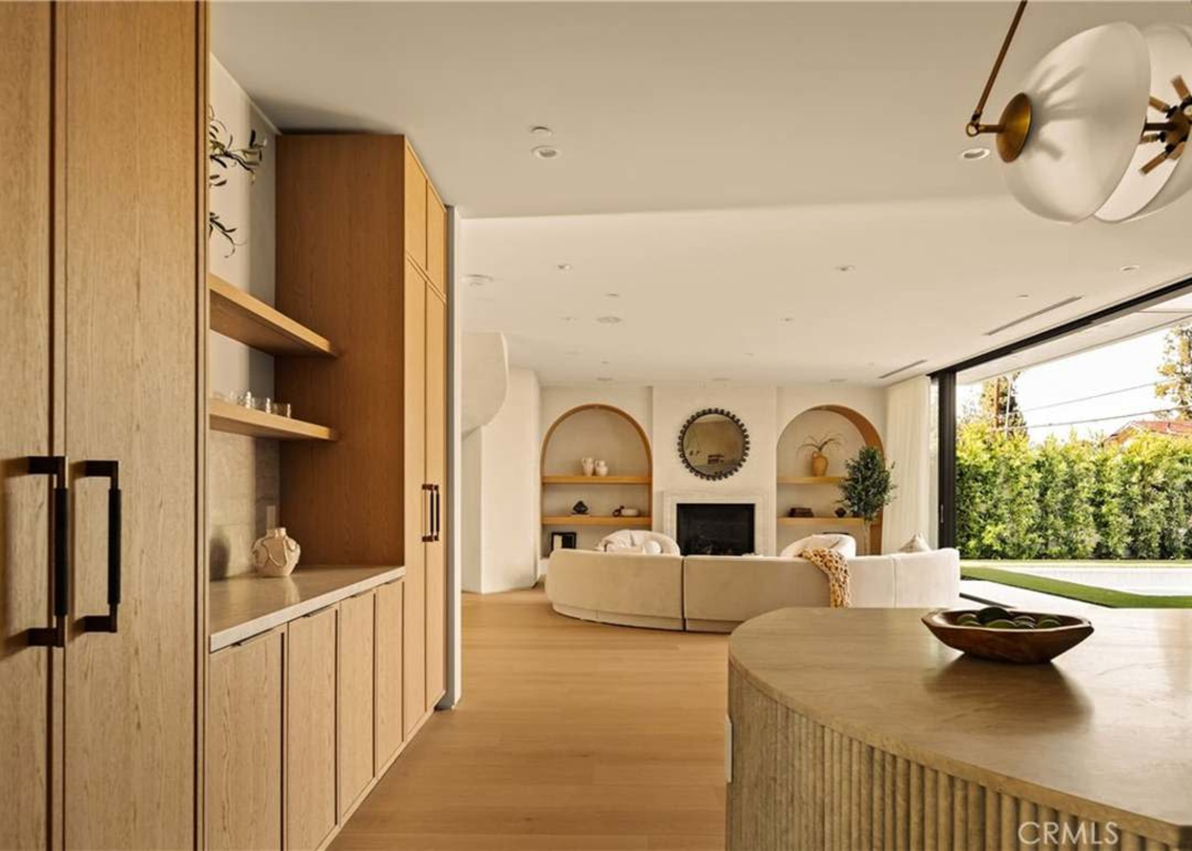 A modern living space features light wooden cabinetry, a circular mirror above a fireplace, and a sectional sofa facing large windows that open to a garden.