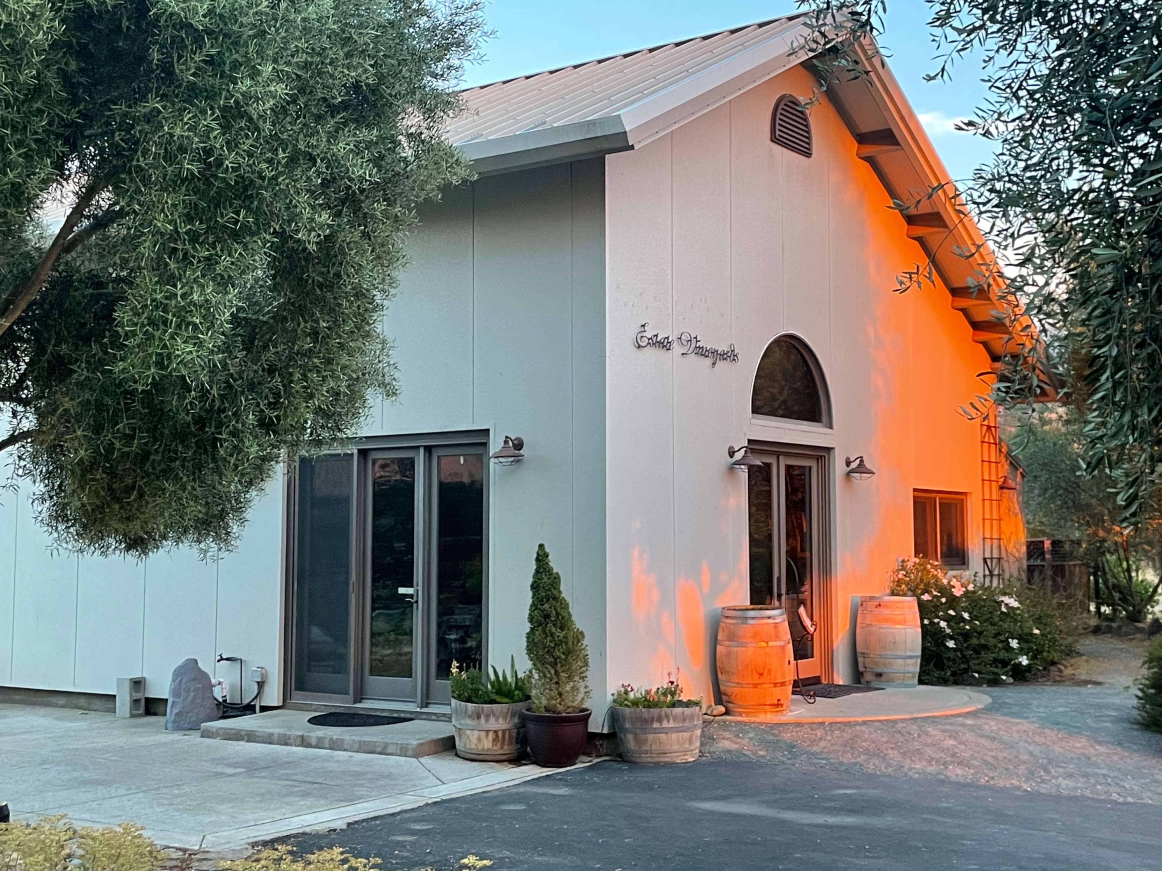 A white, modern building with large windows, a peaked roof, and decorative barrels on either side of the entrance, set amidst greenery.